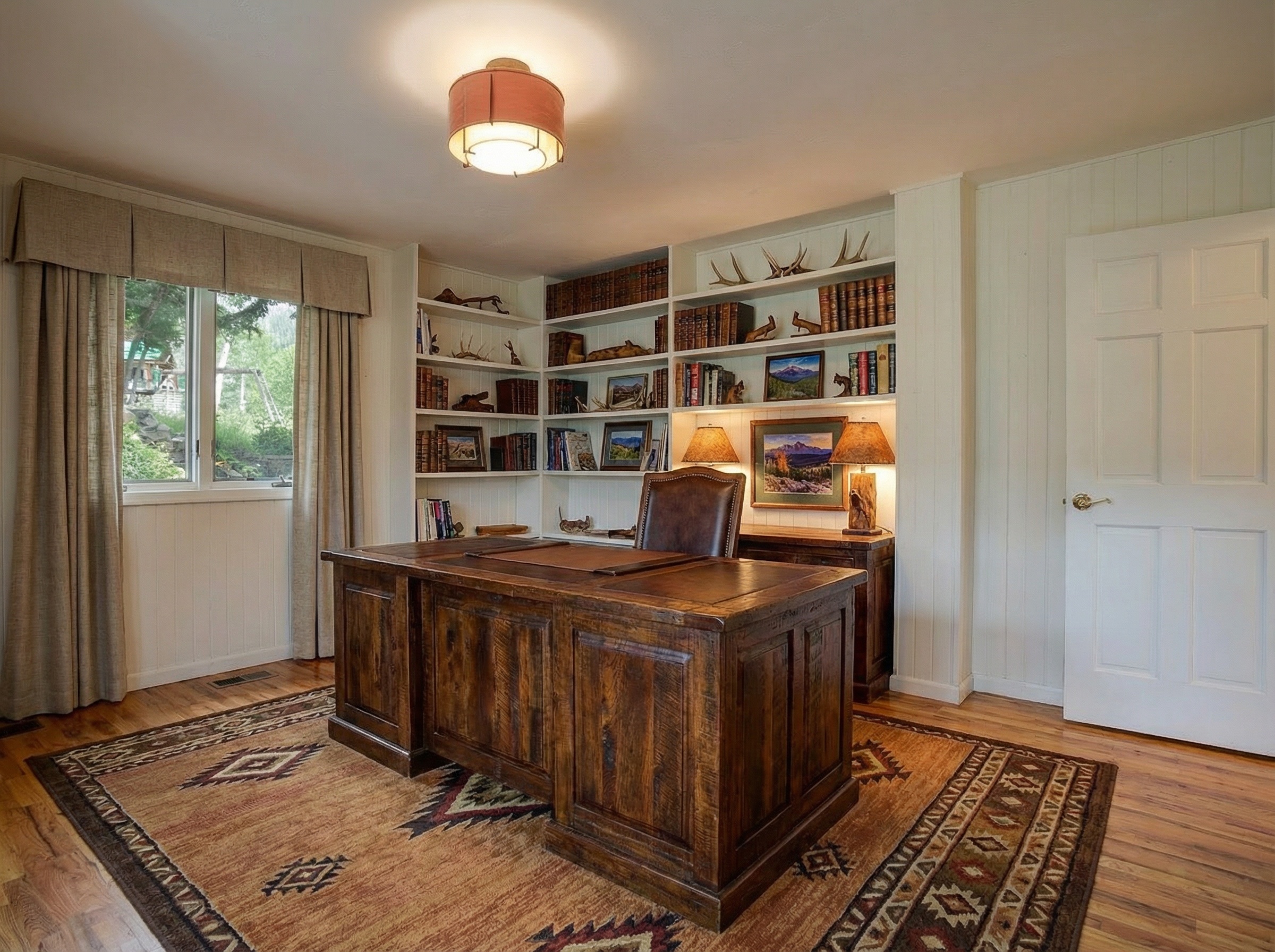 This is an interior shot of a well-appointed home office. The room features a large wooden desk centered on a decorative rug, which contrasts with the wood flooring. Built-in bookshelves filled with books and decorative items line one wall, and a window with neutral-toned curtains allows natural light into the space.