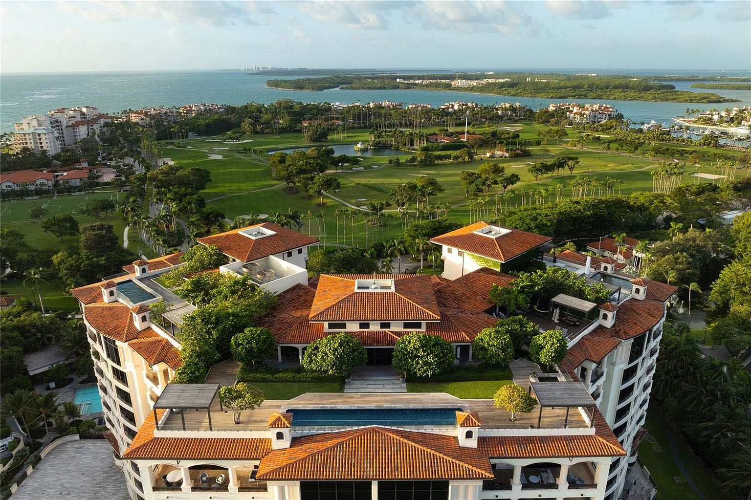 This aerial view presents a luxurious estate featuring a terracotta-tiled roof complex, surrounded by lush greenery and overlooking a meticulously maintained golf course. The architecture showcases a blend of Mediterranean and contemporary design elements. A rooftop pool offers an exclusive amenity with stunning panoramic views of the ocean and neighboring islands, enhancing the property's appeal and setting a high standard for luxury living.