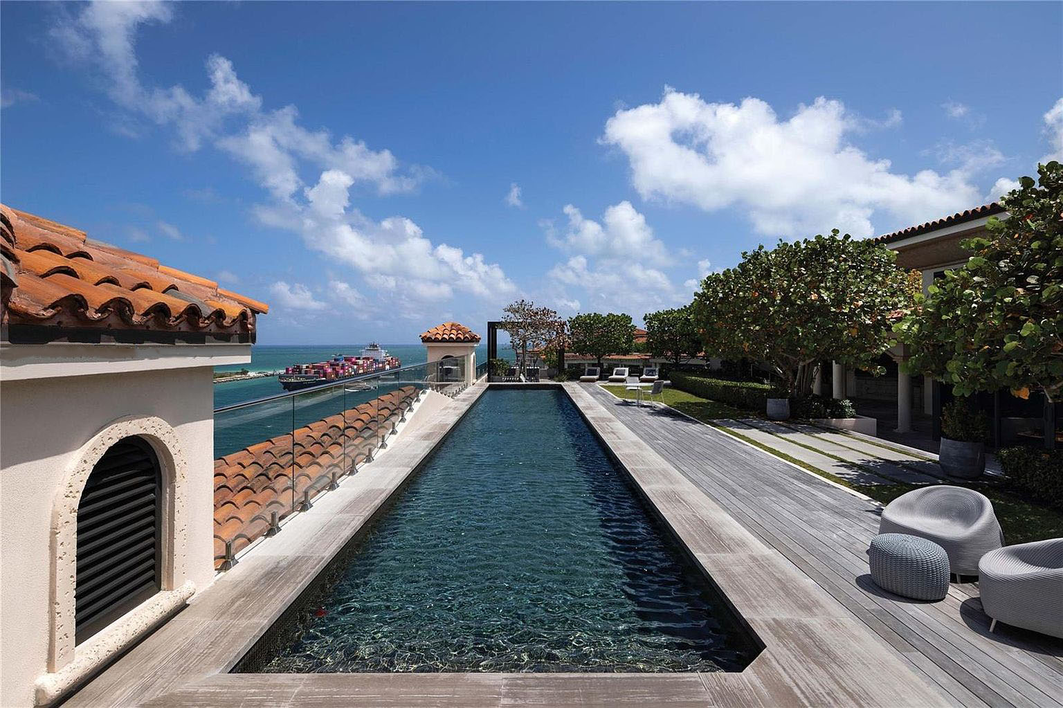 This image showcases an elegant outdoor pool area. The elongated pool is surrounded by a wooden deck and offers stunning views of the ocean with a container ship visible in the distance. The scene combines luxury living with waterfront scenery, enhanced by clear blue skies and scattered clouds.