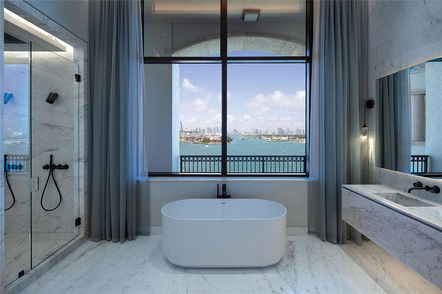 This showcases a luxurious primary bathroom. The room features a modern freestanding bathtub positioned in front of a large window with a city/ocean view, complemented by neutral-toned marble flooring and walls. A glass-enclosed shower and a sleek vanity with dark fixtures complete the spa-like ambiance.