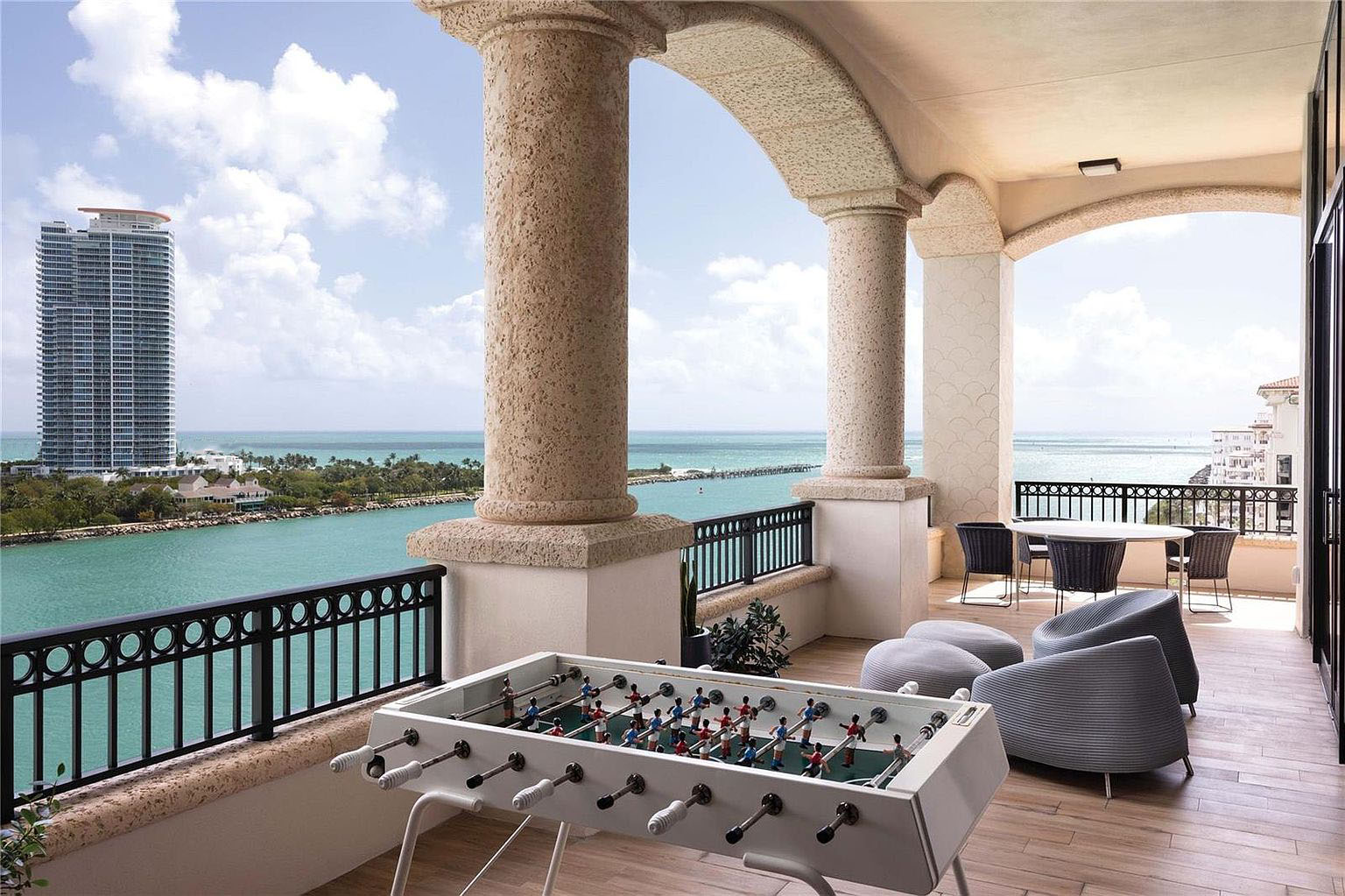 This image showcases a luxurious balcony with striking architectural columns and a foosball table. The balcony is furnished with modern seating and offers a view of the ocean and a high-rise building in the distance. The overall ambiance suggests a blend of leisure and sophisticated outdoor living.