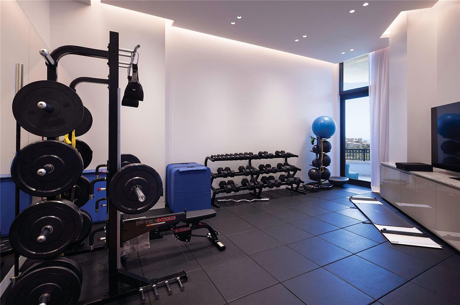 This is a well-equipped home gym featuring a weight rack with numerous plates, various dumbbells on racks, and an exercise ball station. The room has dark, rubber tile flooring and ample natural light from a large window, complemented by recessed ceiling lights. A sleek cabinet and a flat-screen TV add a touch of modern comfort to the workout space.