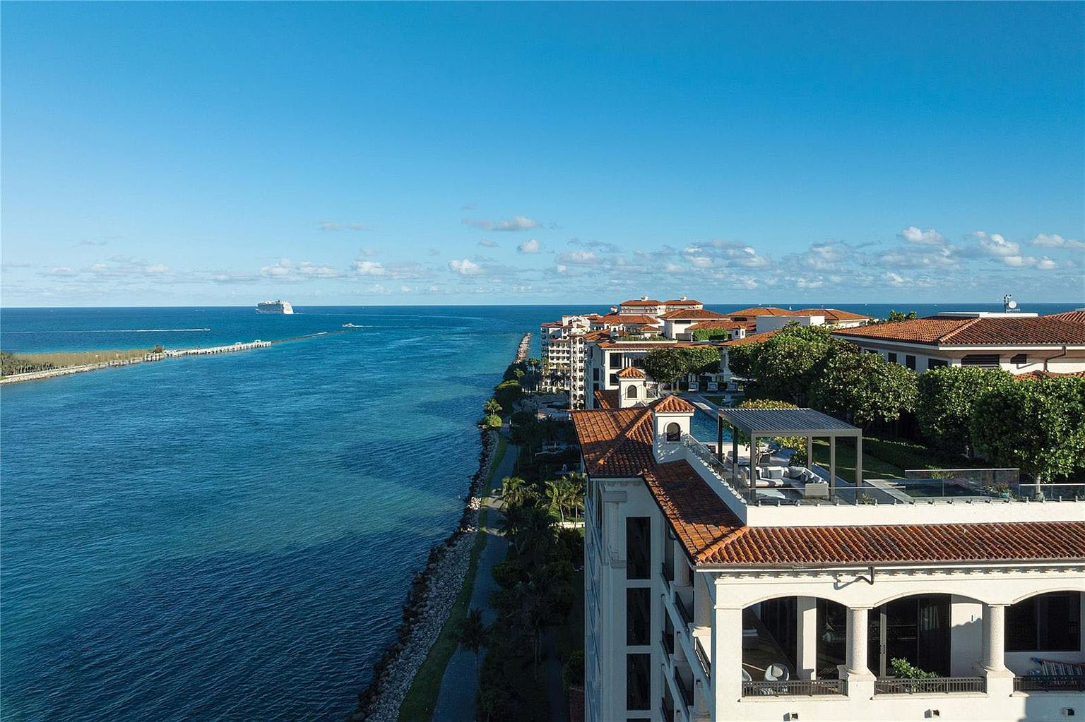This aerial shot showcases a luxury waterfront property featuring Mediterranean-style architecture with red tile roofs. The building boasts multiple levels with balconies and views of the deep blue ocean. Lush landscaping surrounds the property, enhancing its appeal and offering a glimpse of an elevated outdoor lounge.
