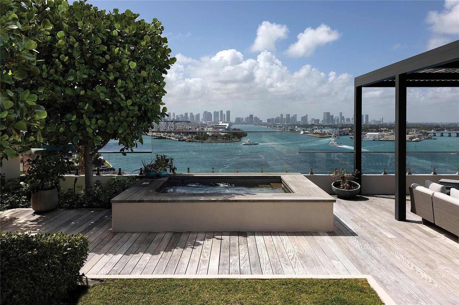 This image showcases a stunning patio or deck with a luxurious spa or hot tub overlooking a breathtaking cityscape and waterfront. The area is elegantly designed with wood decking, a pergola or shaded structure, and tasteful landscaping. The overall impression is one of upscale outdoor living and relaxation, enhancing the property's appeal for entertaining and enjoying the views.