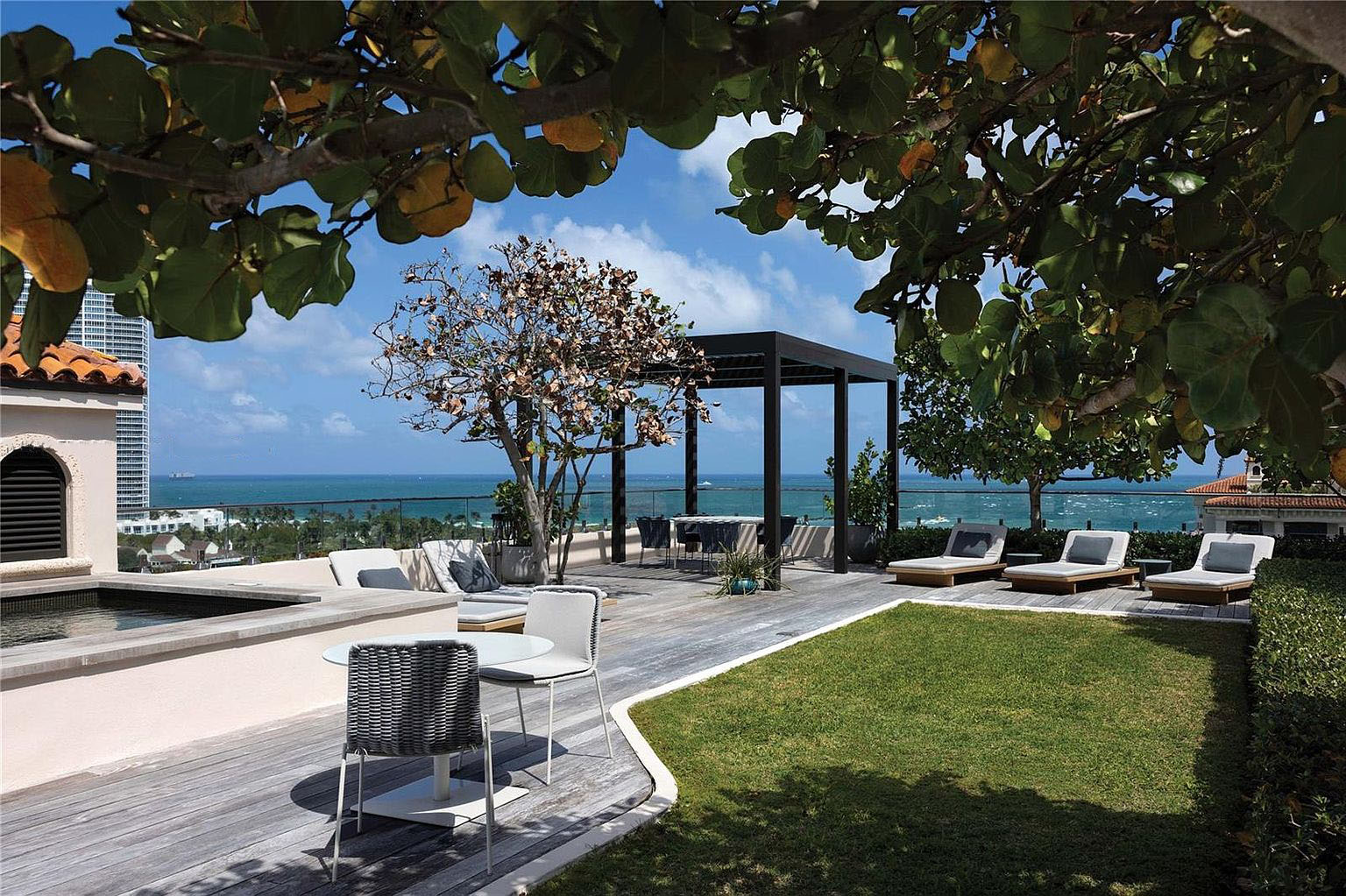 This image showcases a luxurious rooftop patio/deck/balcony, designed for relaxation and entertainment. The space features modern furniture, a pergola, and a well-maintained lawn area, all with stunning ocean views. The design incorporates both hardscaping and landscaping, creating a sophisticated outdoor living area perfect for enjoying the coastal ambiance.