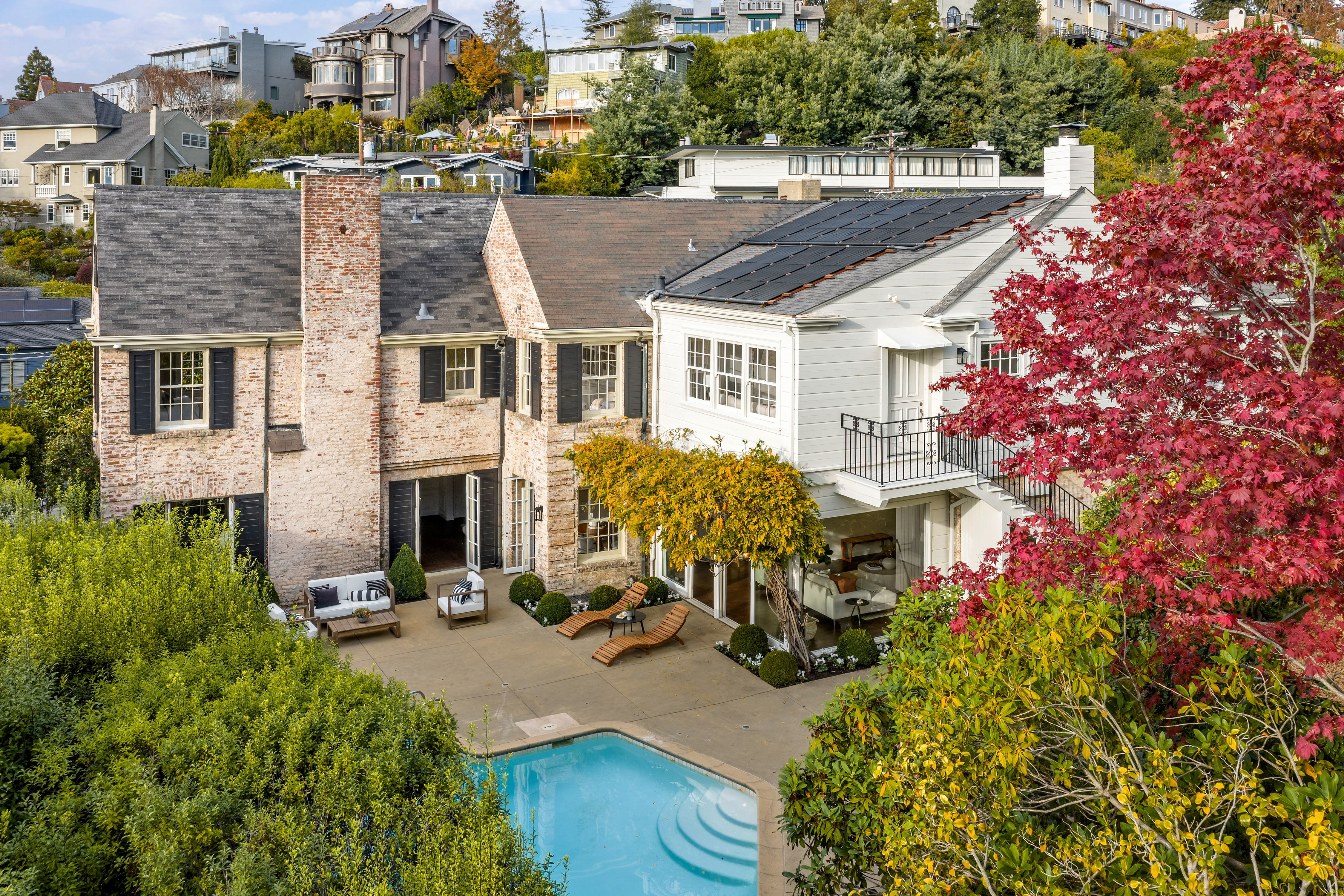 This aerial view showcases a luxurious home with a brick facade, a well-maintained backyard featuring a swimming pool, and a patio area with lounge furniture. The property also includes solar panels on the roof and is surrounded by lush greenery, offering a sense of privacy and tranquility. The perspective is high-angle, providing a comprehensive view of the property and its surroundings.