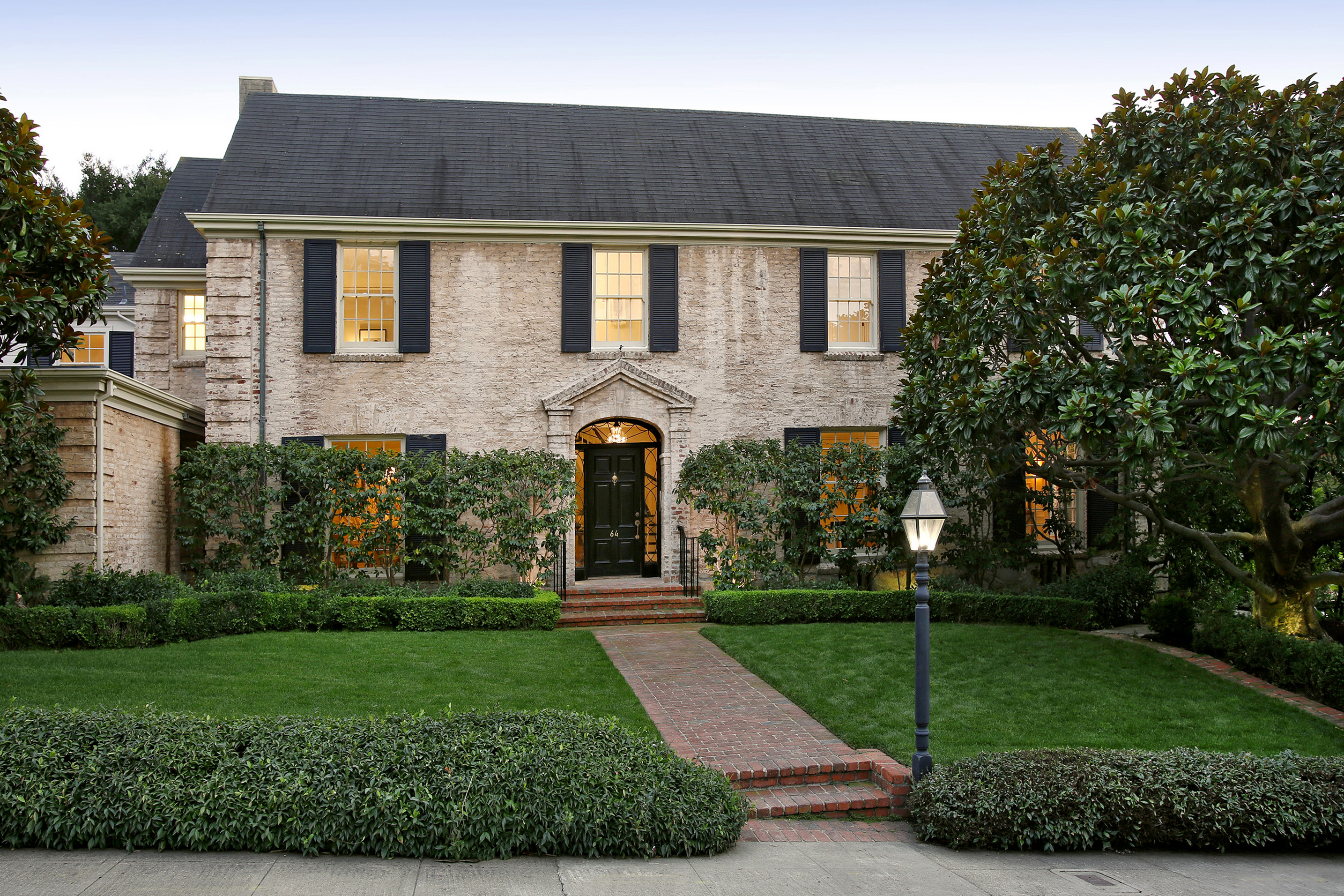 This is a stately two-story brick home with a well-manicured lawn and mature landscaping. The house features black shutters, a dark-colored roof, and a brick walkway leading to the front door. A classic lamppost adds to the curb appeal, creating an inviting and elegant first impression.