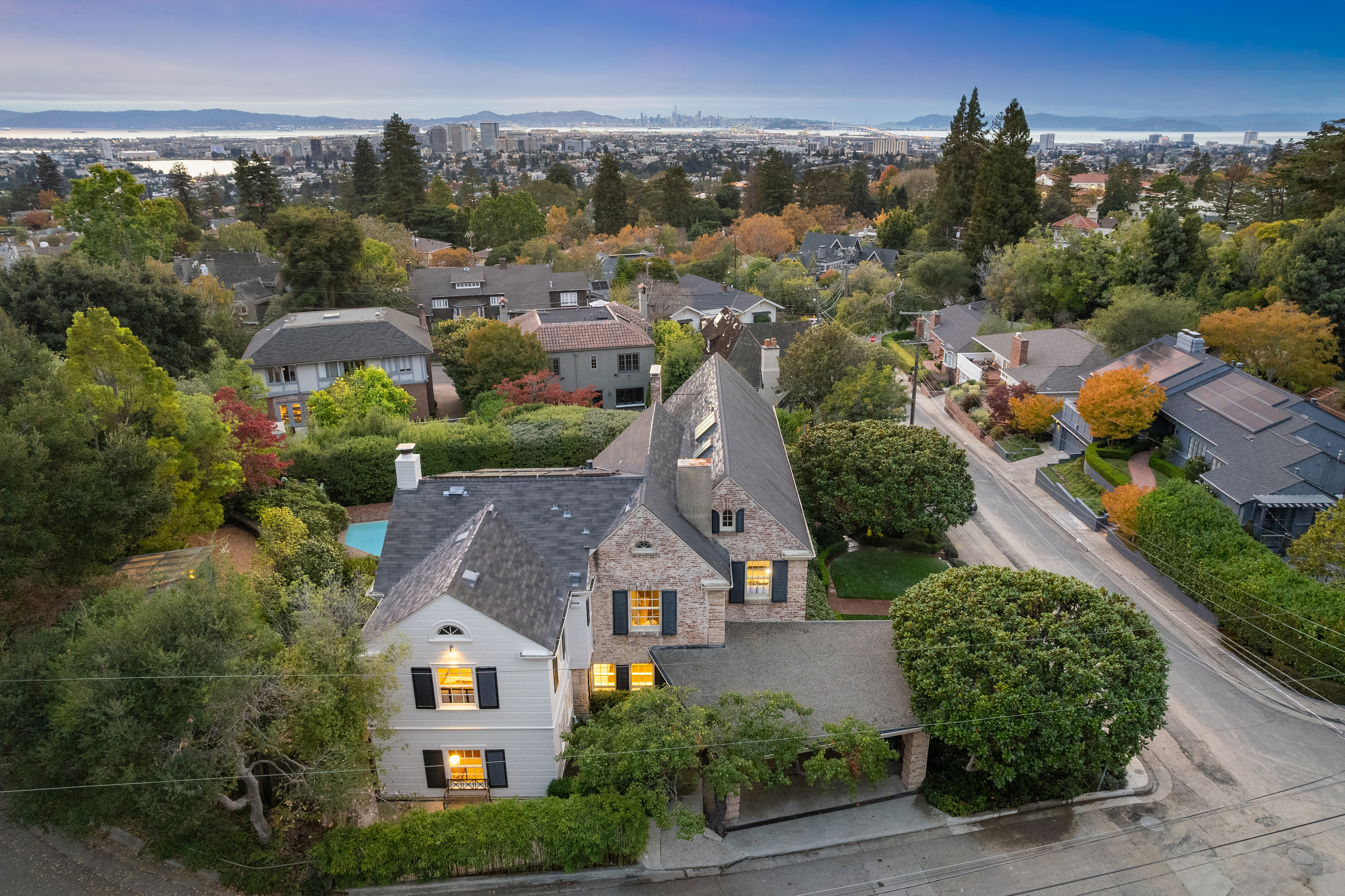 This aerial view showcases a large, multi-story home with a combination of stone and white siding, complemented by a dark gray roof. The property is surrounded by lush greenery and mature trees, offering a sense of privacy and seclusion. In the background, a cityscape is visible, suggesting a desirable location with both natural beauty and urban convenience.
