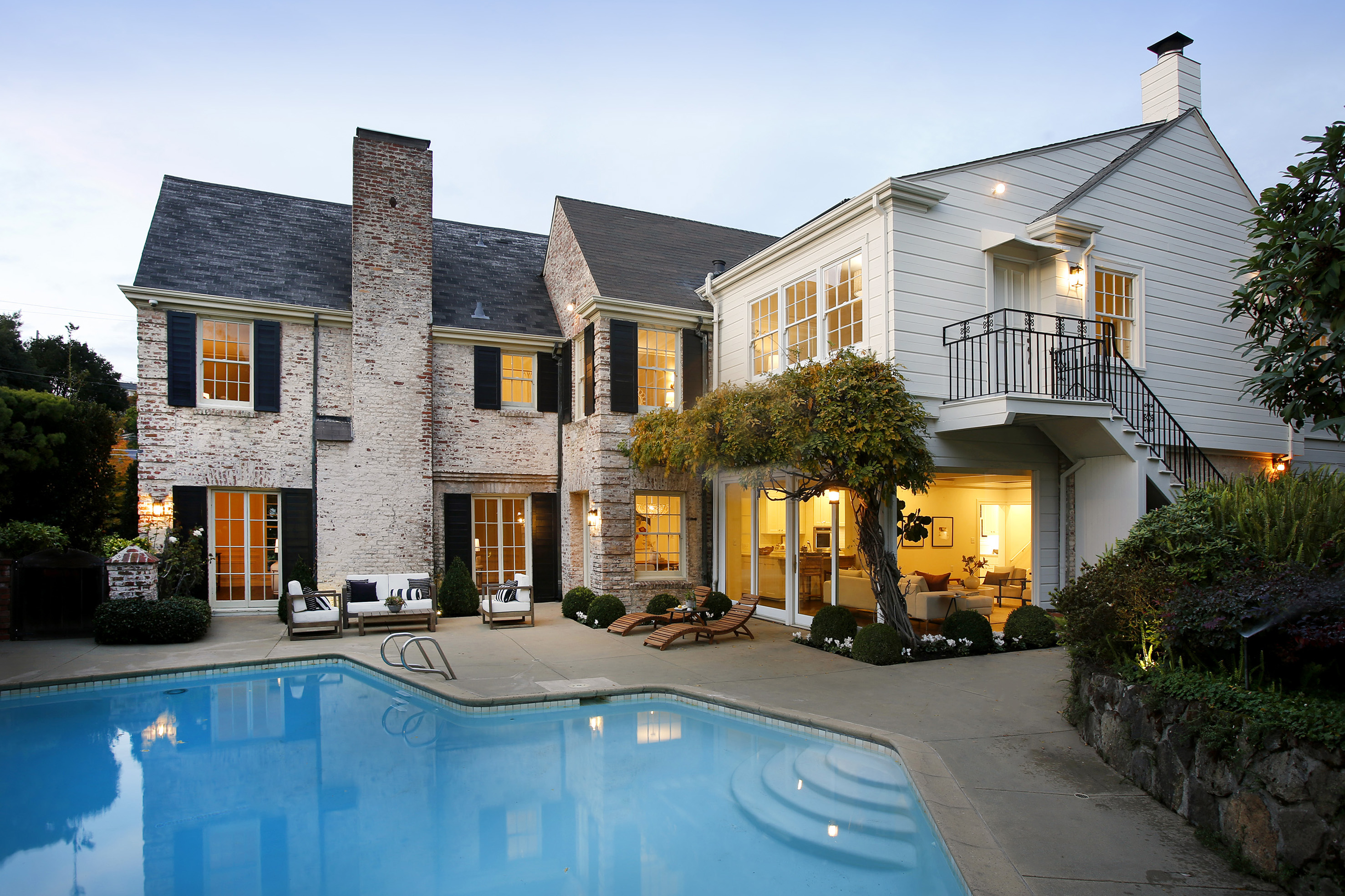 This image showcases the rear exterior of a luxurious home, highlighting a stunning swimming pool as the focal point. The house features a blend of brick and white siding, with multiple windows and a balcony overlooking the pool area. Outdoor furniture is arranged on the patio, creating an inviting space for relaxation and entertainment.