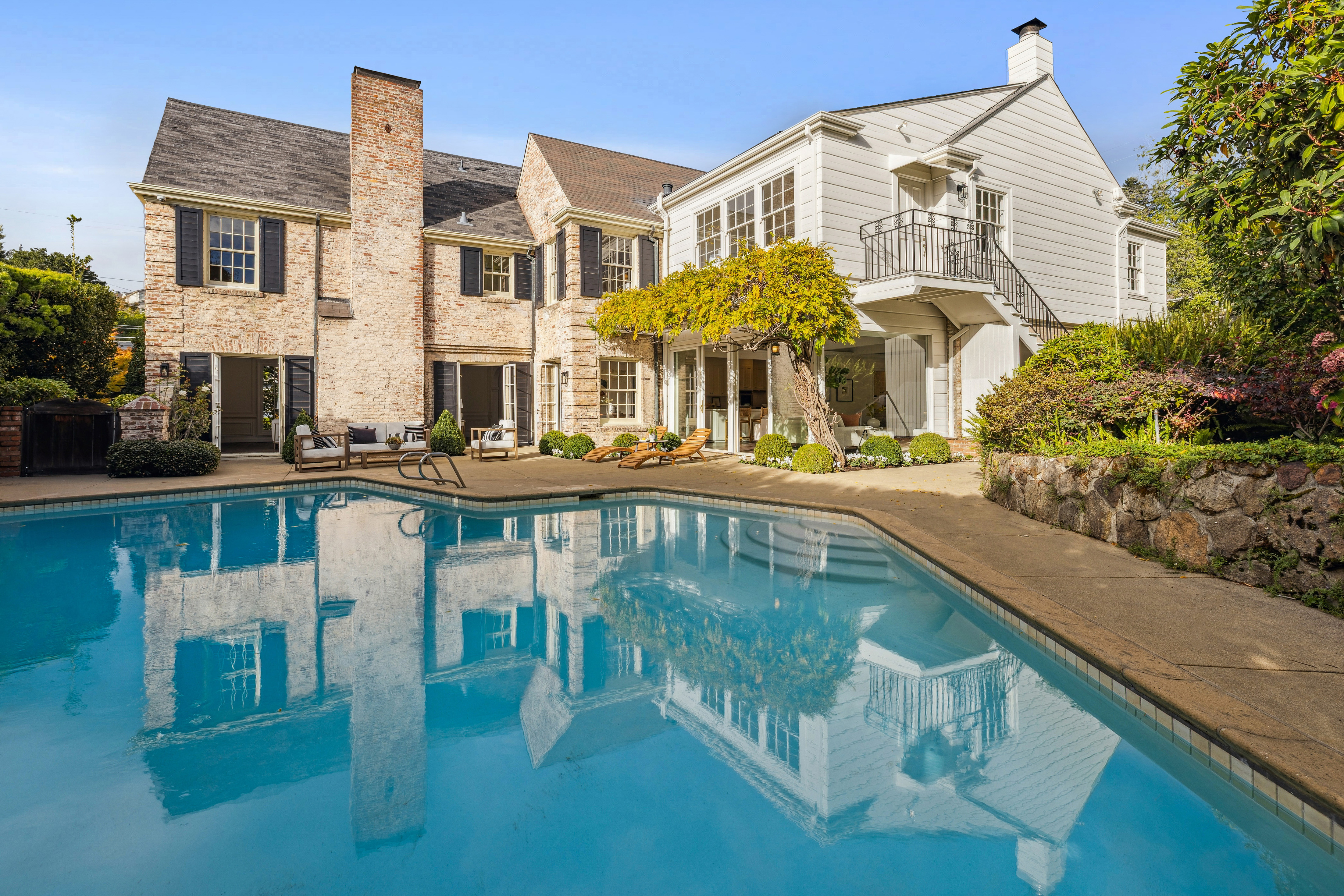 This image showcases the rear exterior of a luxurious home, highlighting a pristine swimming pool that reflects the architecture. The property features a blend of brick and white siding, complemented by well-manicured landscaping and a charming balcony. The scene evokes a sense of tranquility and upscale living, perfect for relaxation and entertainment.