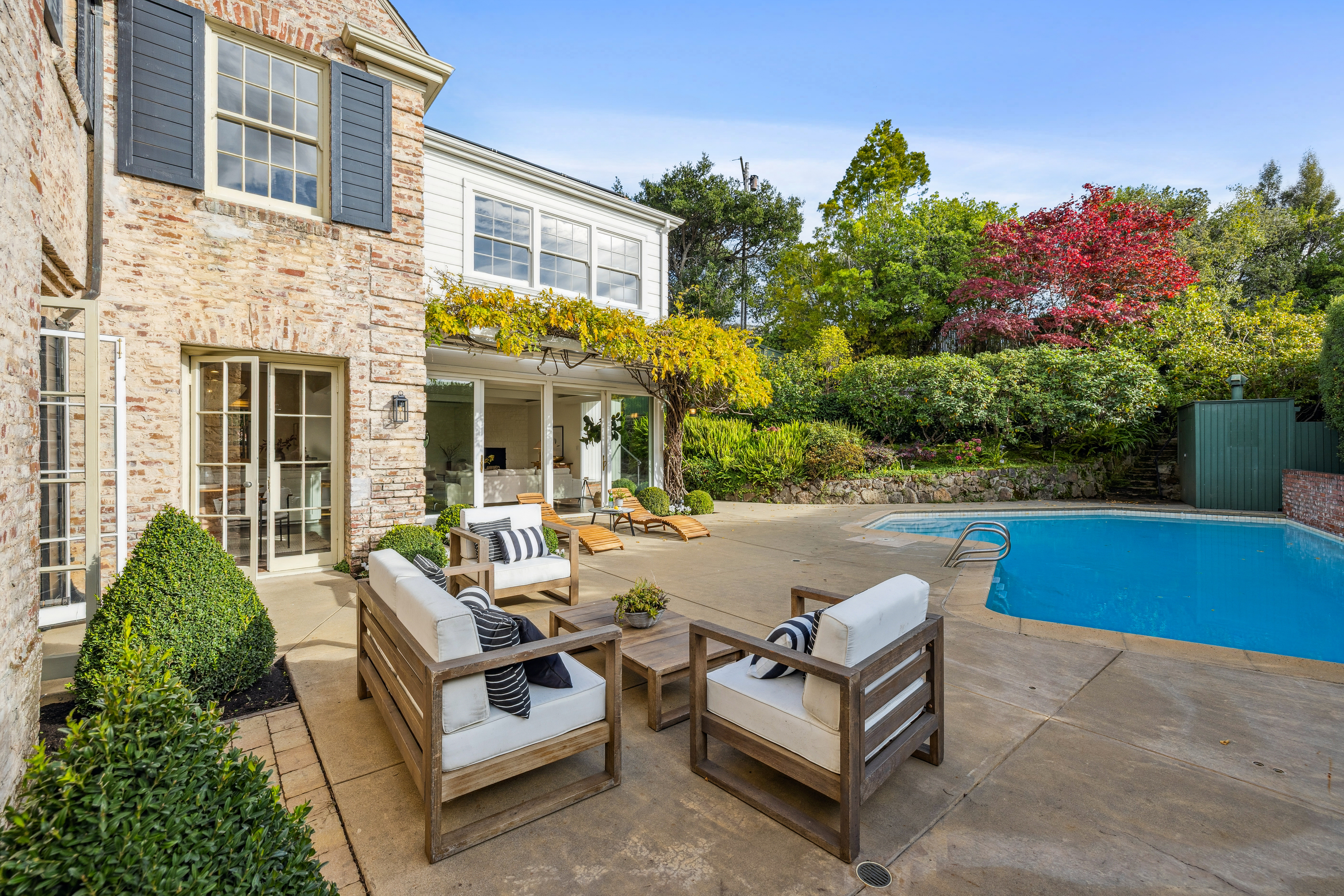 This is a view of the backyard featuring a pool and patio area. The patio is furnished with outdoor seating, including chairs and loungers. The pool is surrounded by lush landscaping, including trees and shrubs, creating a private and inviting outdoor space.
