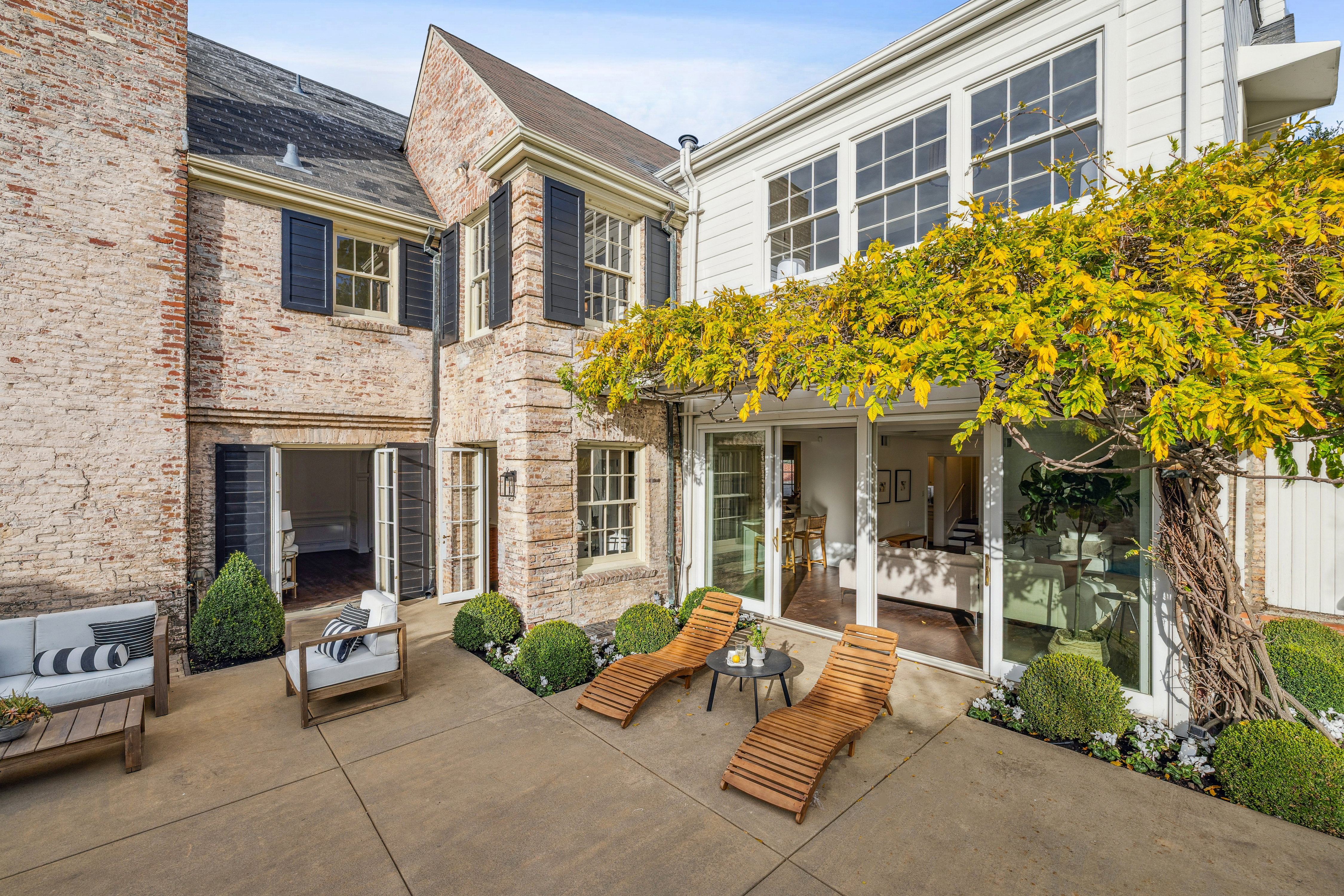 This image showcases a charming patio area, featuring comfortable outdoor seating arrangements including a sofa, chair, and two lounge chairs. The patio is surrounded by lush greenery and a vine-covered pergola, creating a serene and inviting outdoor living space. The architecture combines brick and white siding, adding to the property's character and appeal.