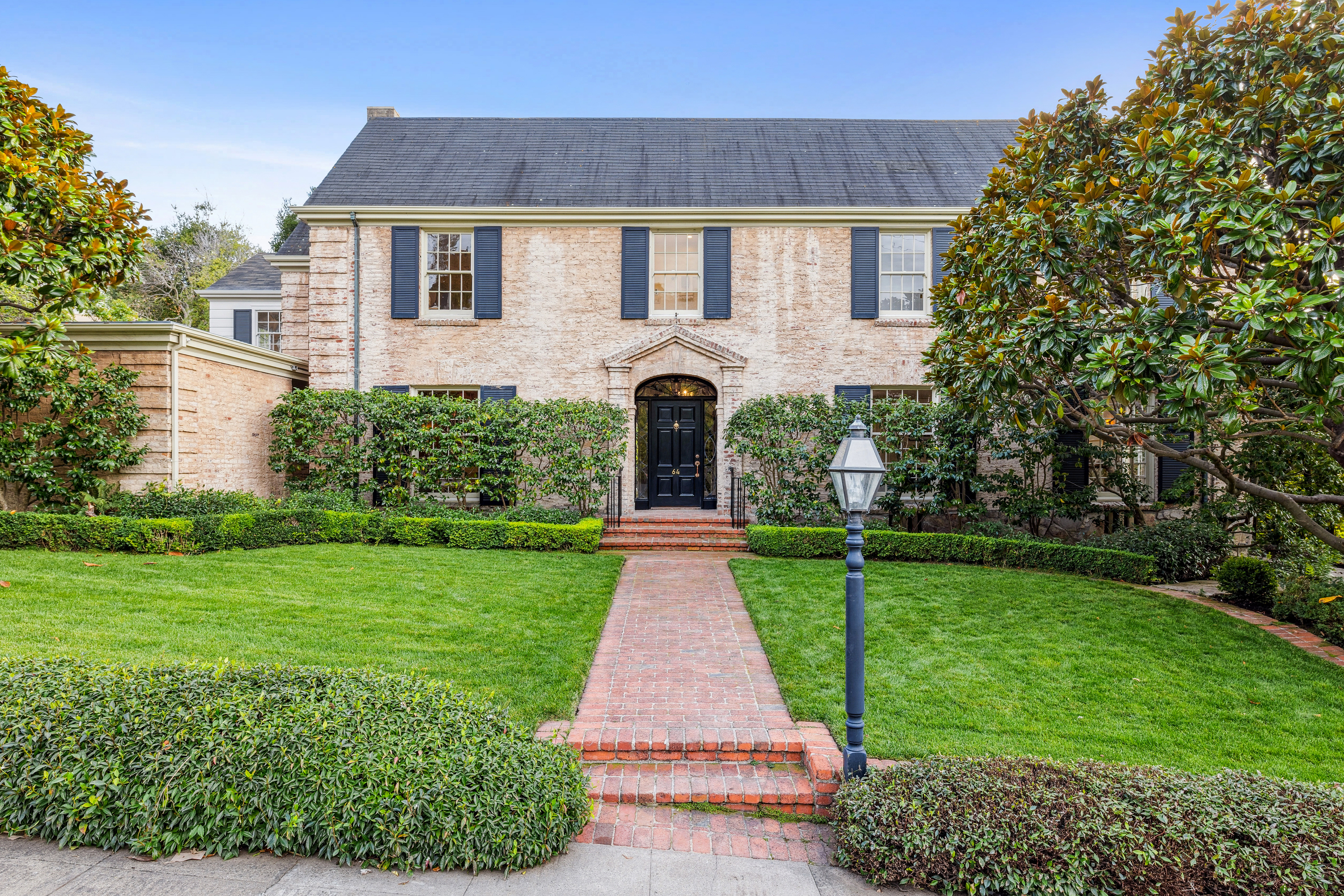 This is a stately two-story brick home with a well-manicured lawn and mature landscaping. The house features dark blue shutters, a black front door with an arched entryway, and a brick walkway leading to the entrance. The overall impression is one of classic elegance and curb appeal.