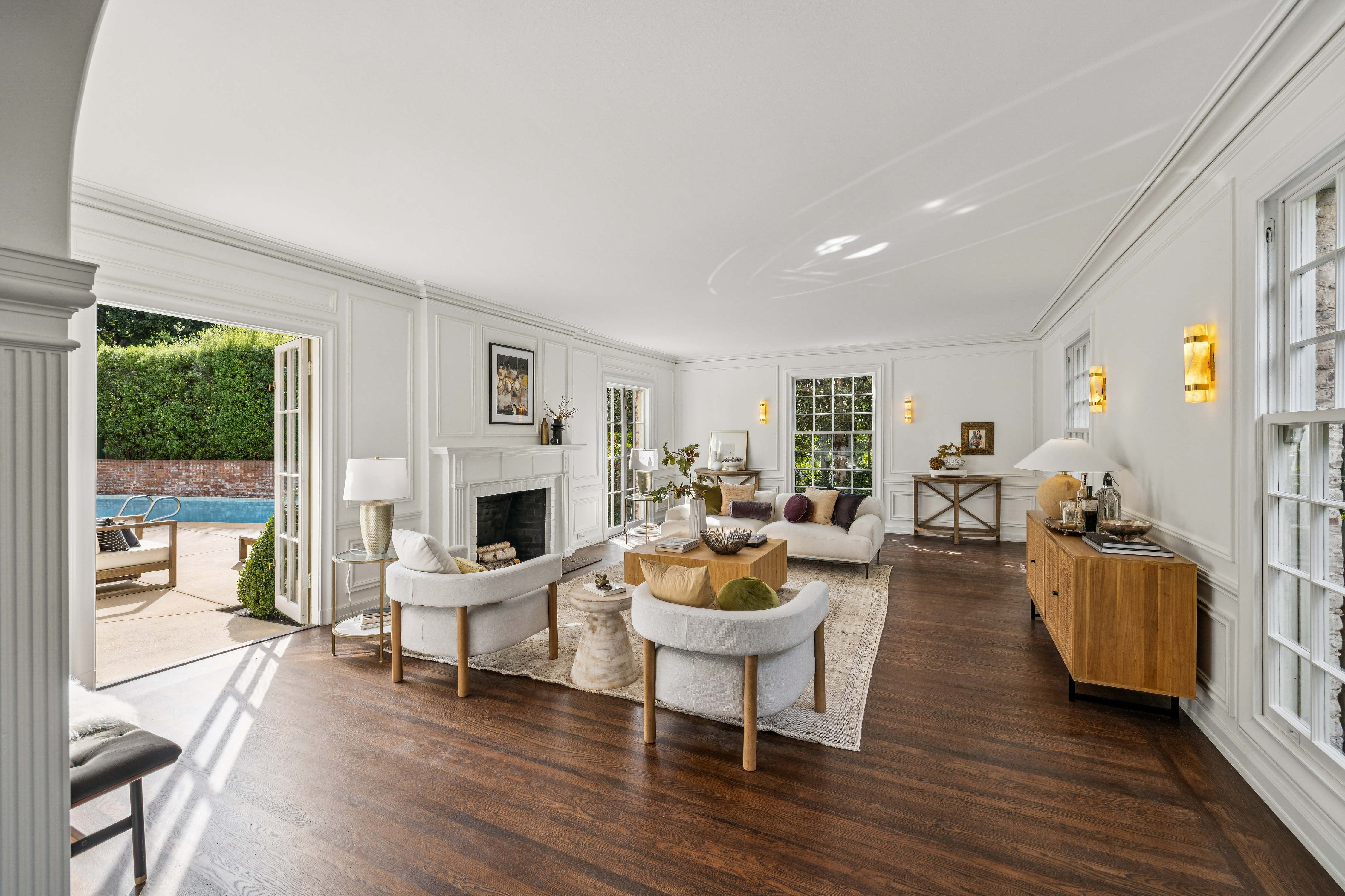 This is a bright and spacious living room featuring hardwood floors, white walls with detailed molding, and large windows that provide ample natural light. The room is furnished with a neutral-toned sofa, two armchairs, a fireplace, and a wooden console table, creating a comfortable and elegant atmosphere. French doors open to an outdoor patio and pool area, seamlessly blending indoor and outdoor living.