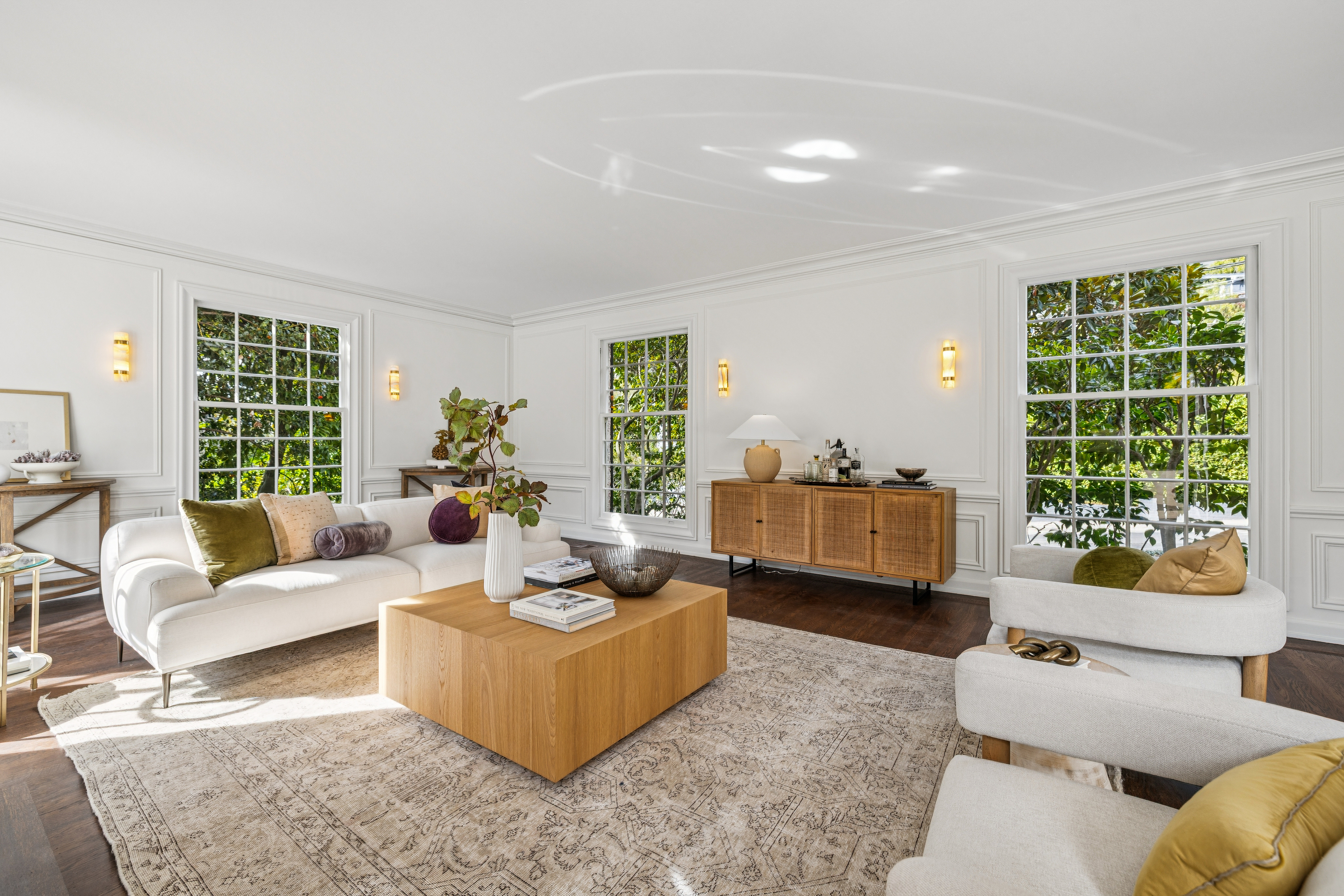 This is a bright and airy living room featuring a white sofa with decorative pillows, a large wooden coffee table, and two armchairs. The room is well-lit with natural light streaming through large windows, complemented by elegant wall sconces. A woven sideboard adds texture and warmth to the space, while a patterned rug anchors the seating arrangement.