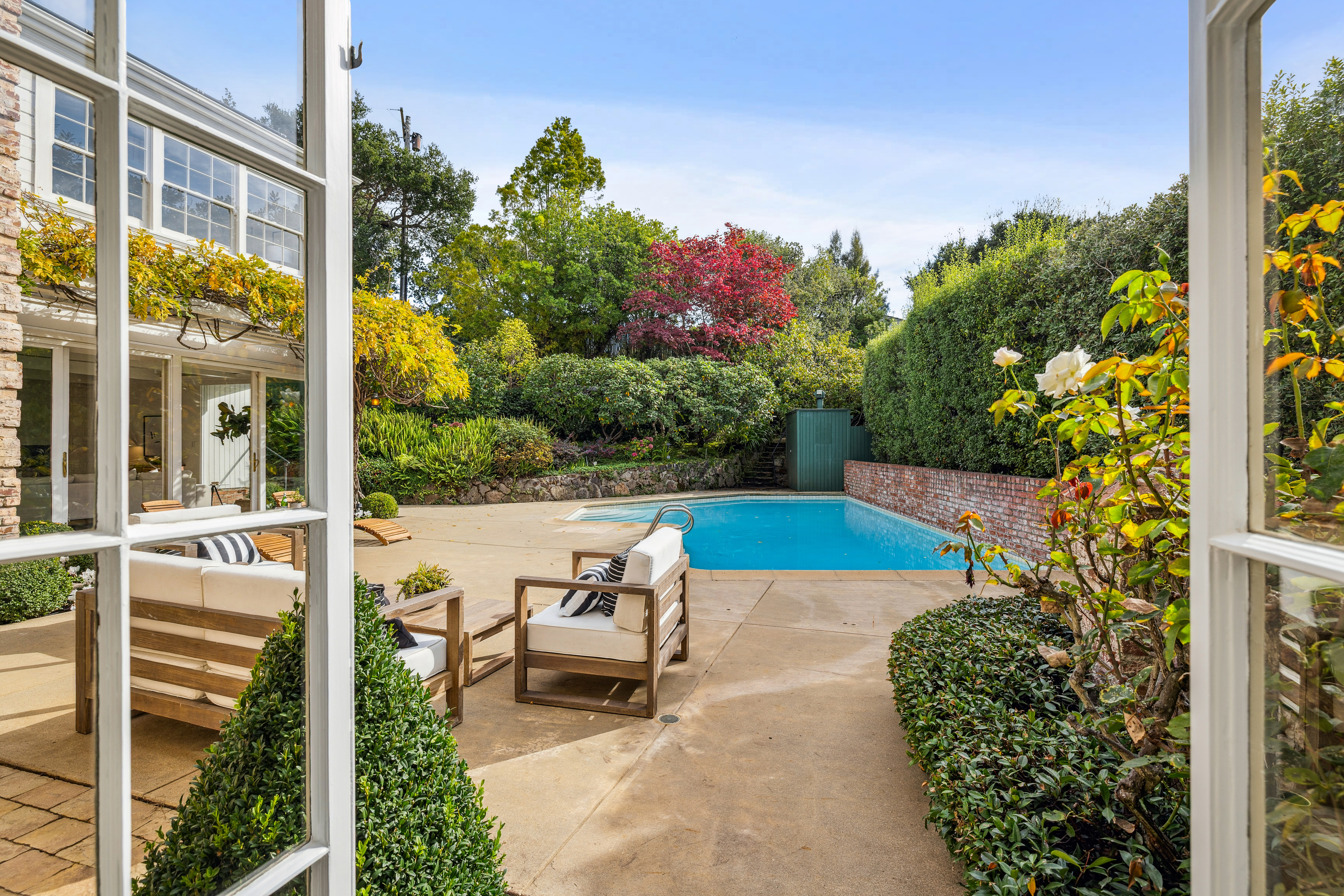 This image showcases a serene backyard pool area, viewed through an open window. The pool is surrounded by a concrete patio with outdoor seating, lush greenery, and mature trees, creating a private and inviting oasis. The scene evokes a sense of relaxation and luxury, perfect for entertaining or unwinding.