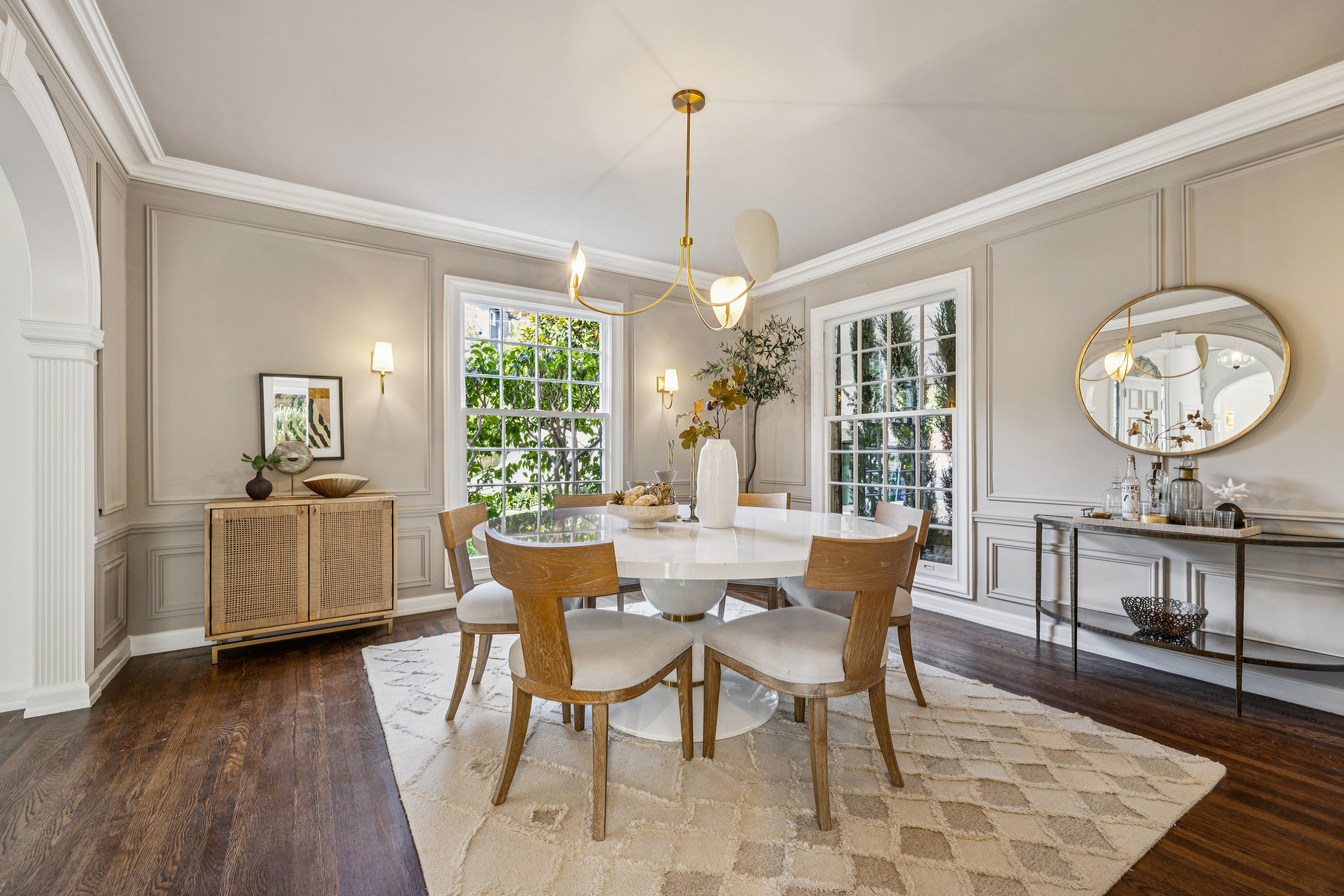This is an interior shot of a dining room featuring a round white table surrounded by six wooden chairs with light-colored cushions. A modern chandelier hangs above the table, and natural light floods the room through two large windows. A sideboard and a console table with a round mirror add to the room's elegance, all set on a patterned area rug over dark wood floors.