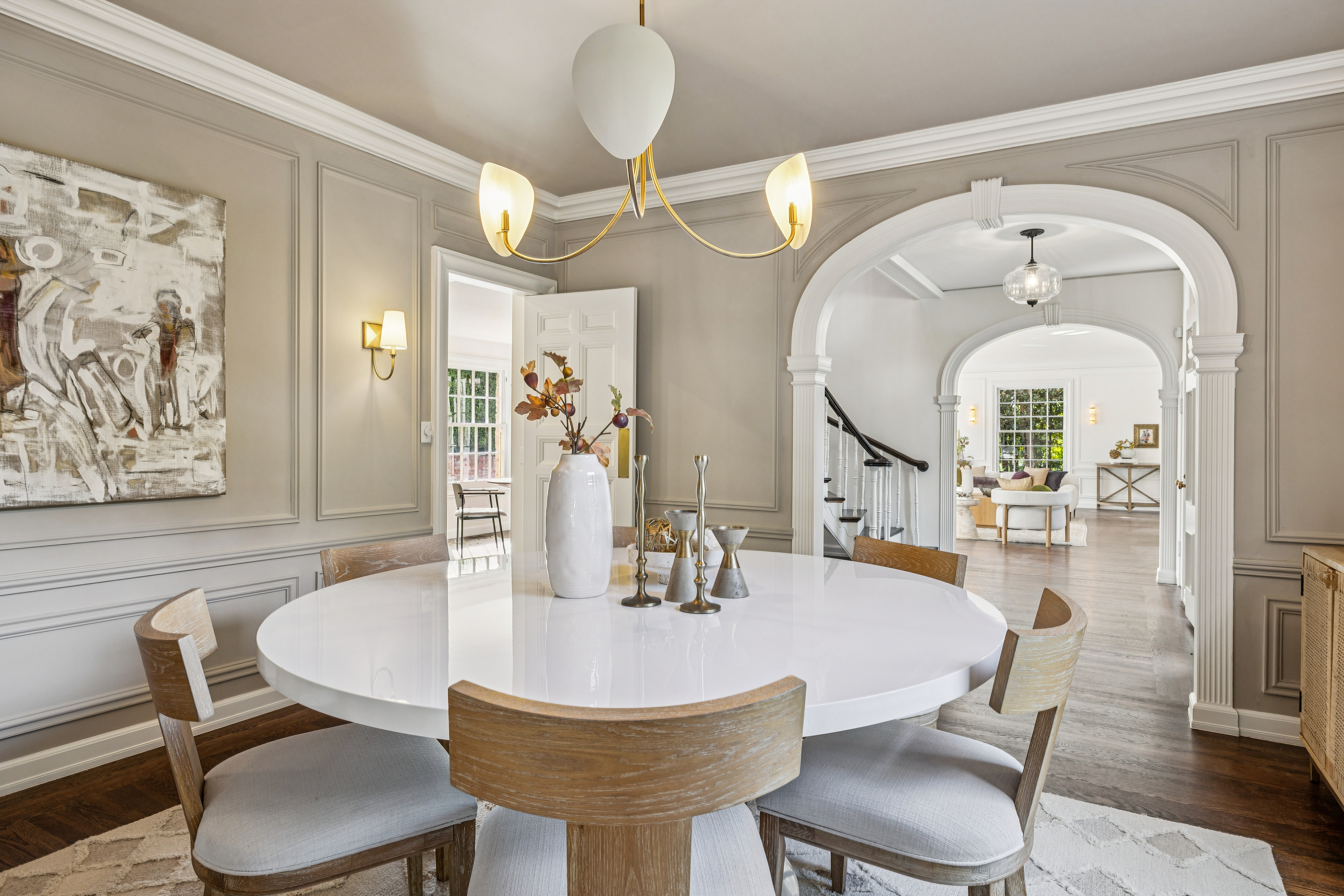 This is an interior shot of a dining room featuring a large, round, white table surrounded by chairs with light wood frames and light-colored upholstery. A modern chandelier hangs above the table, and artwork adorns the walls. The room opens into a hallway with an arched doorway, providing a glimpse into other areas of the home. The perspective is from a medium distance, showcasing the entire dining area and its connection to the rest of the house.