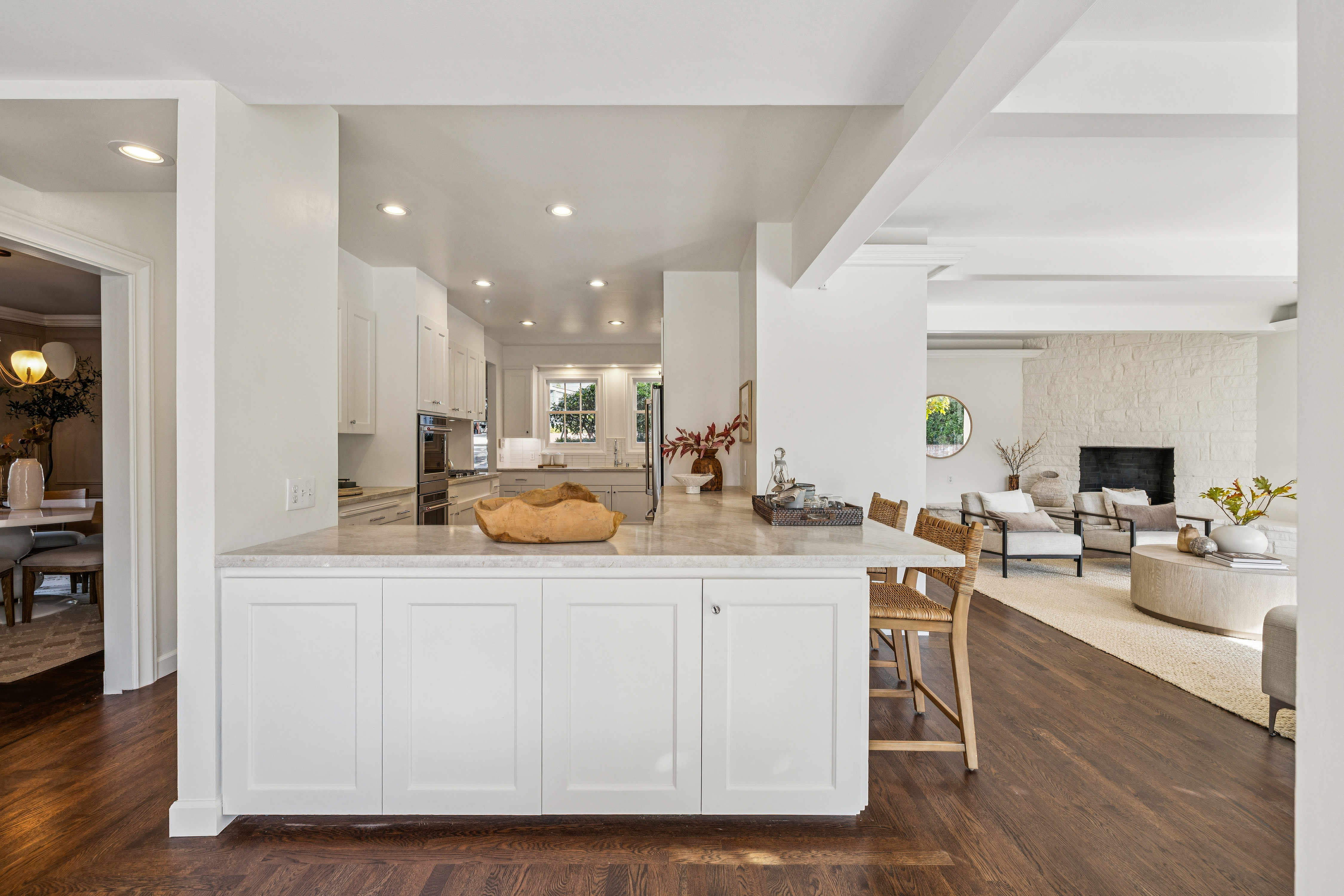 This interior shot showcases a bright, open kitchen that seamlessly transitions into a living area. The kitchen features white cabinetry, a marble countertop island with bar seating, and modern appliances. The living area boasts a stone fireplace and comfortable seating, creating an inviting and cohesive living space.