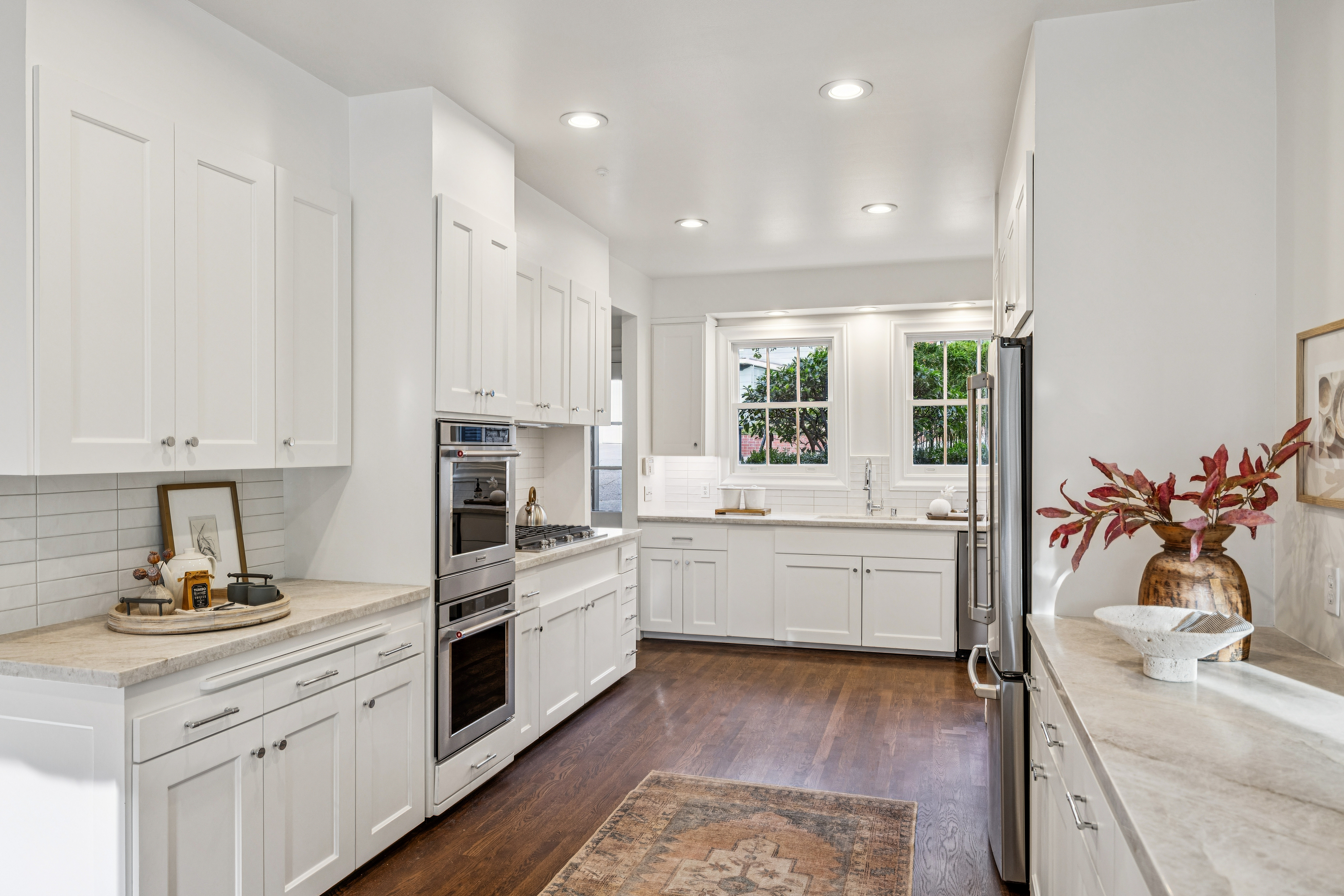 This is a bright and modern kitchen featuring white cabinetry, stainless steel appliances, and light-colored countertops. The kitchen is well-lit with recessed lighting and natural light from the windows. The dark hardwood floors and area rug add warmth to the space, creating an inviting atmosphere.
