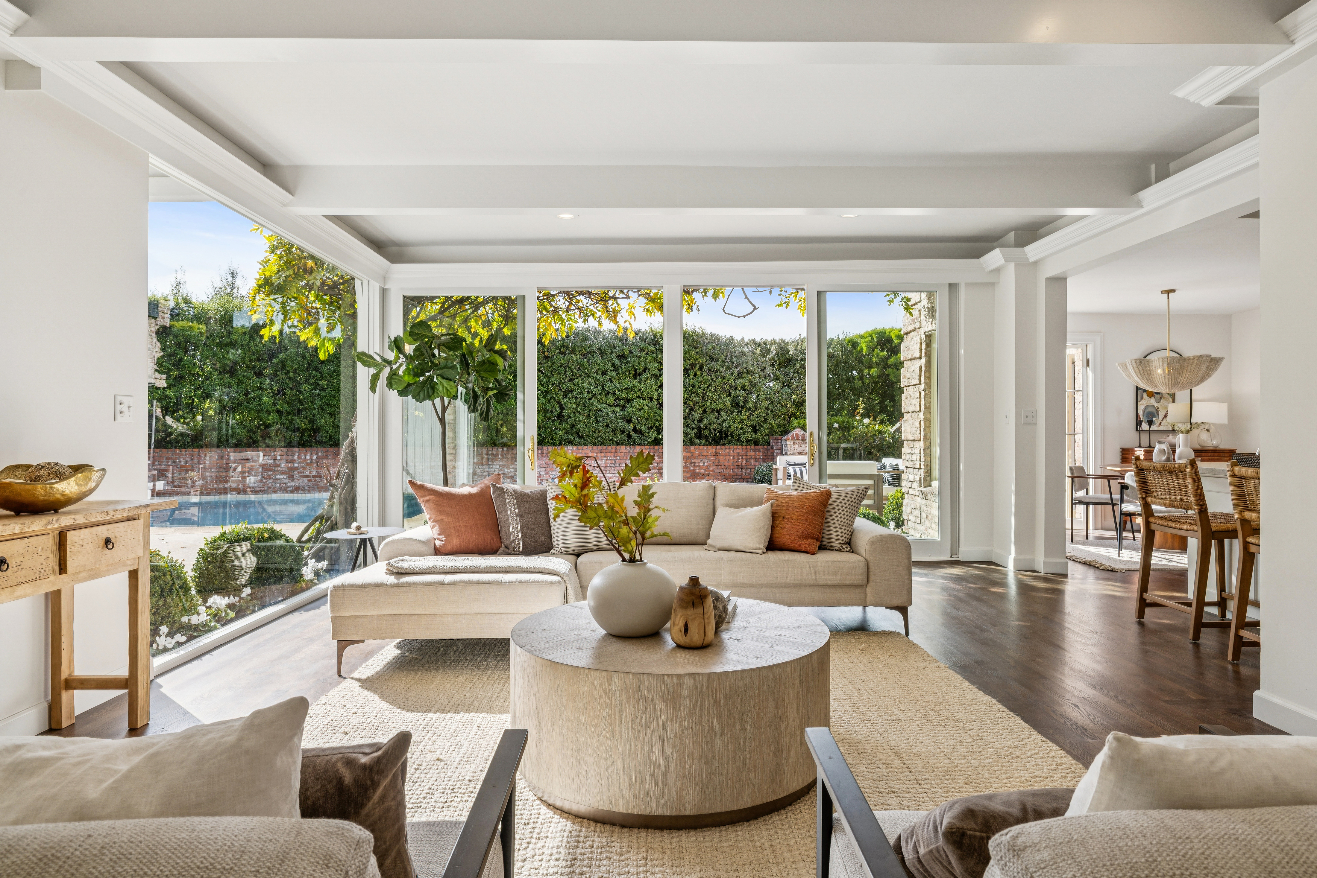 This is a bright and airy living room featuring a large sectional sofa with decorative pillows, a round coffee table with decorative items, and a neutral-toned area rug. Large windows offer views of the outdoor greenery and pool, creating a seamless indoor-outdoor living experience. The room is well-lit and appears spacious, with a glimpse into the dining area.