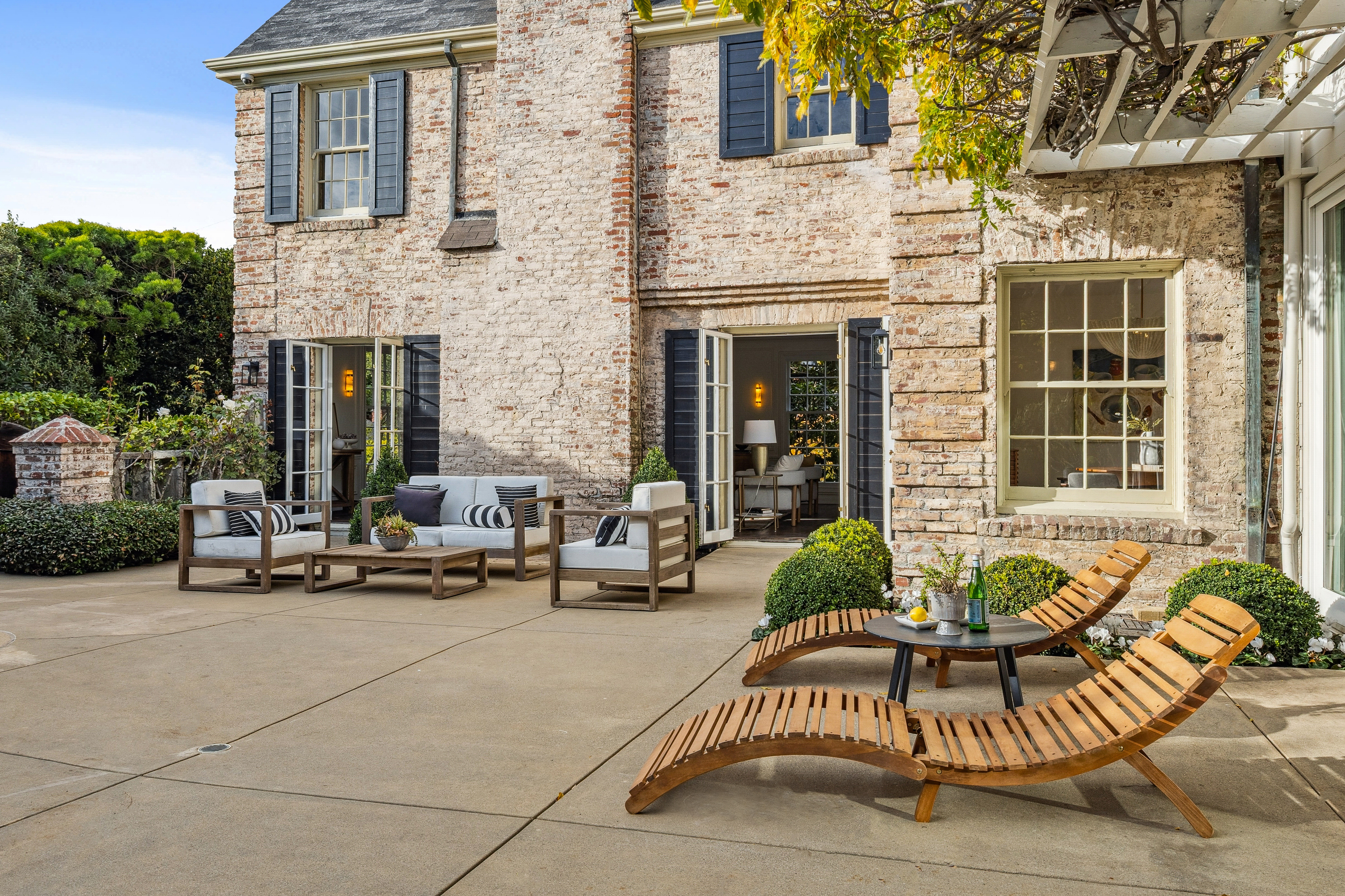 This image showcases a charming patio area with a mix of lounge seating and wooden chaise lounges, perfect for relaxation and entertaining. The patio is adjacent to a brick building with dark shutters and open doors, offering a seamless transition between indoor and outdoor living. Lush greenery surrounds the space, enhancing the sense of privacy and tranquility.