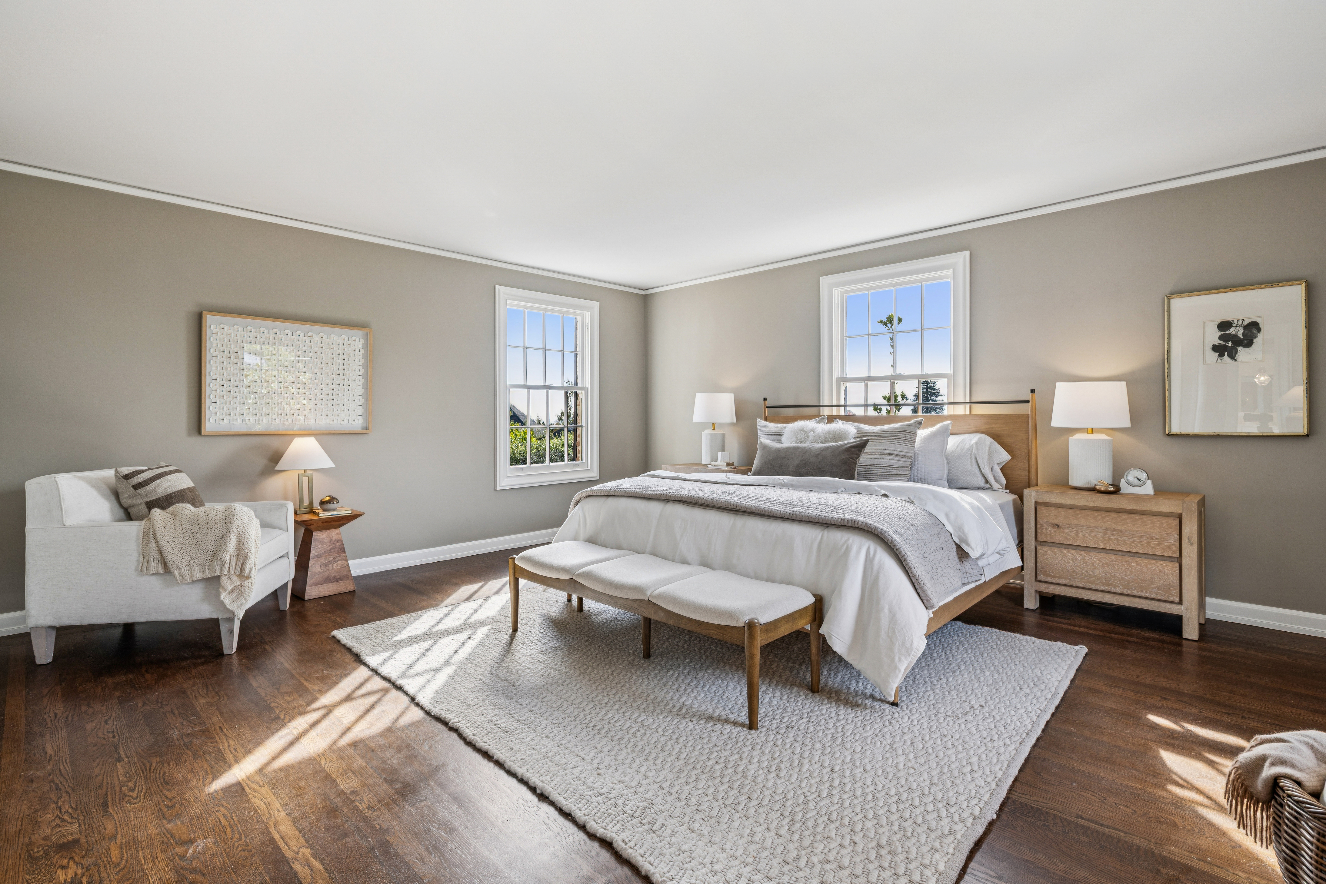 This is a bright and airy primary bedroom featuring hardwood floors, neutral-toned walls, and two large windows that provide ample natural light. The room is centered around a wooden-framed bed with white linens and a gray throw, complemented by matching nightstands and lamps. A light-colored area rug anchors the space, adding warmth and texture to the room.