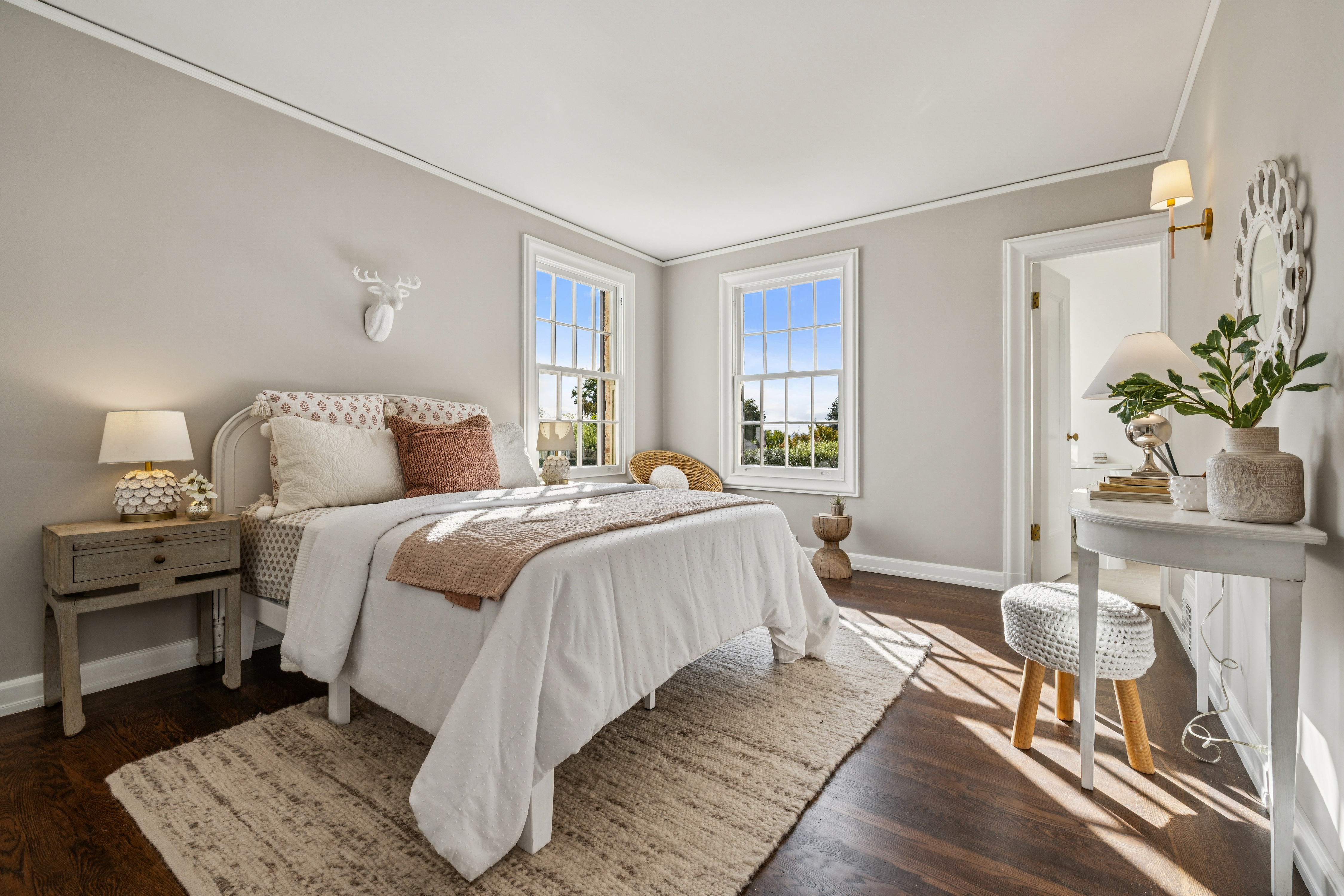This is a bright and airy primary bedroom featuring hardwood floors, two large windows, and neutral wall colors. The room is furnished with a bed, a nightstand, and a vanity table with a stool. The overall style is cozy and inviting, creating a relaxing atmosphere.