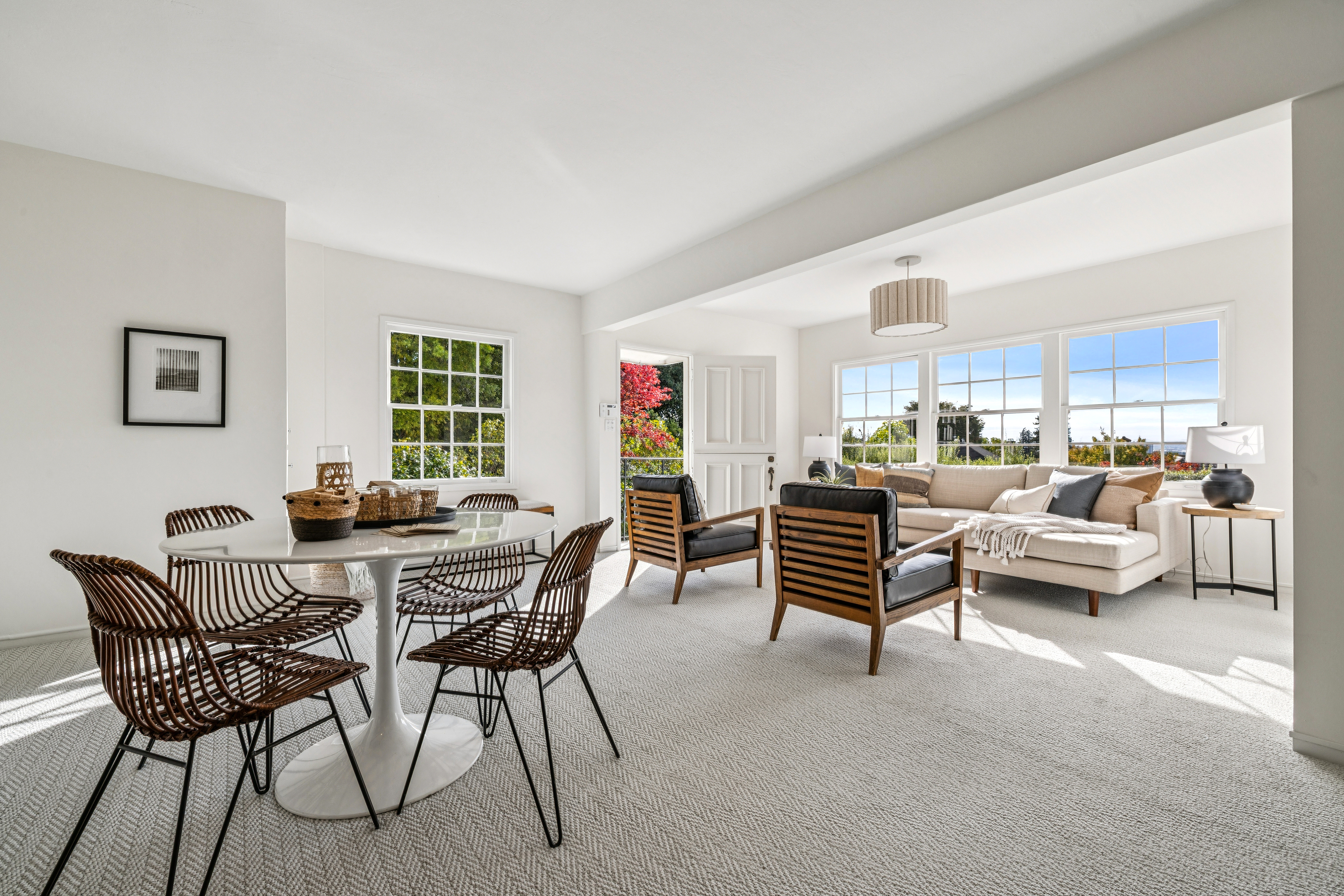 This is an interior shot of a bright and airy living room and dining area. The living room features a neutral-toned sectional sofa, two modern armchairs with wooden frames, and large windows offering natural light. The dining area includes a round white table surrounded by stylish wicker chairs, creating a cohesive and inviting space.