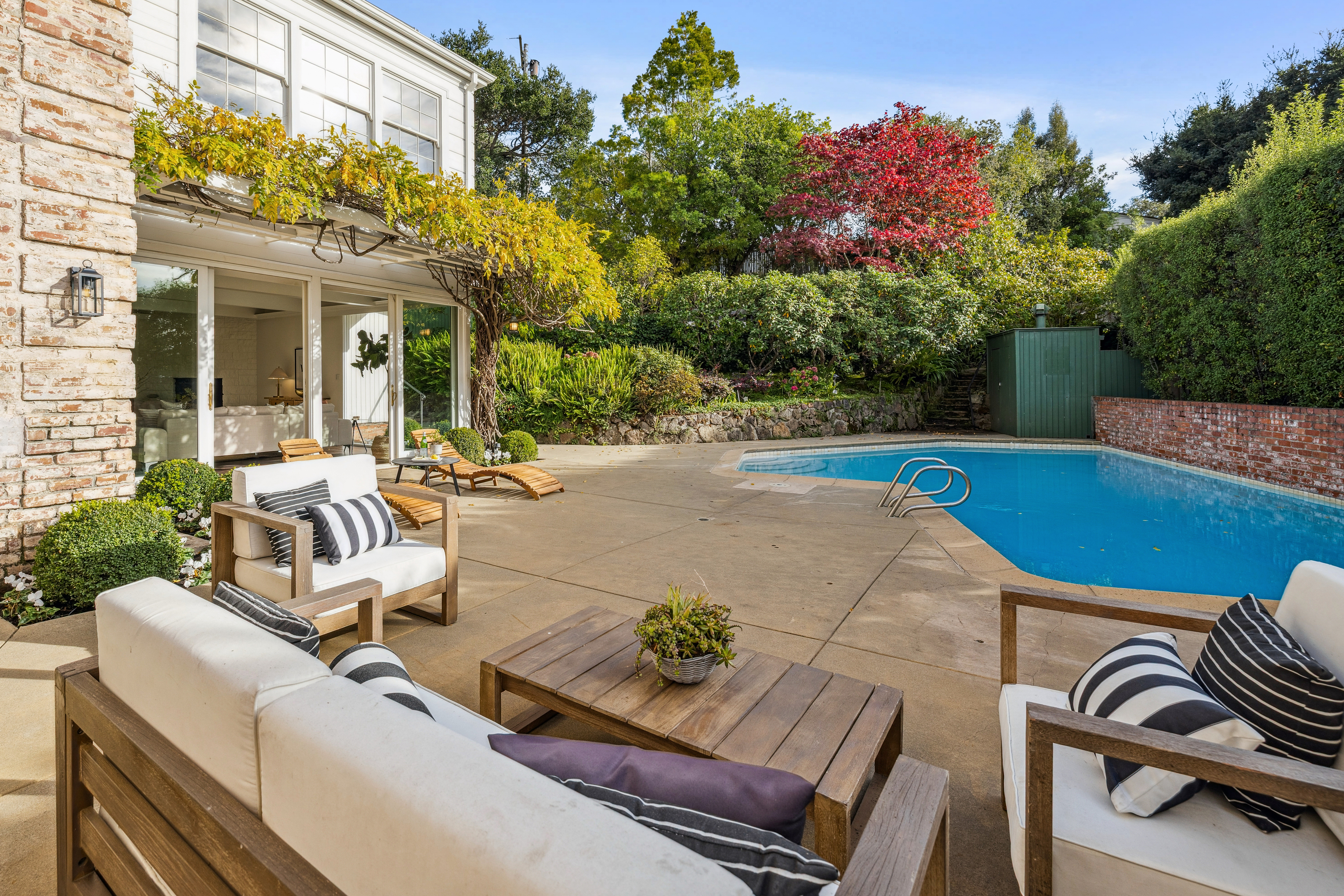 This image showcases a beautiful backyard pool area, featuring a rectangular pool with a stainless steel ladder and a surrounding concrete patio. Outdoor furniture, including a sofa and chairs with striped cushions, are arranged around a wooden coffee table, creating a relaxing lounge space. Lush greenery and trees provide privacy and a vibrant backdrop, enhancing the overall appeal of the outdoor living area.