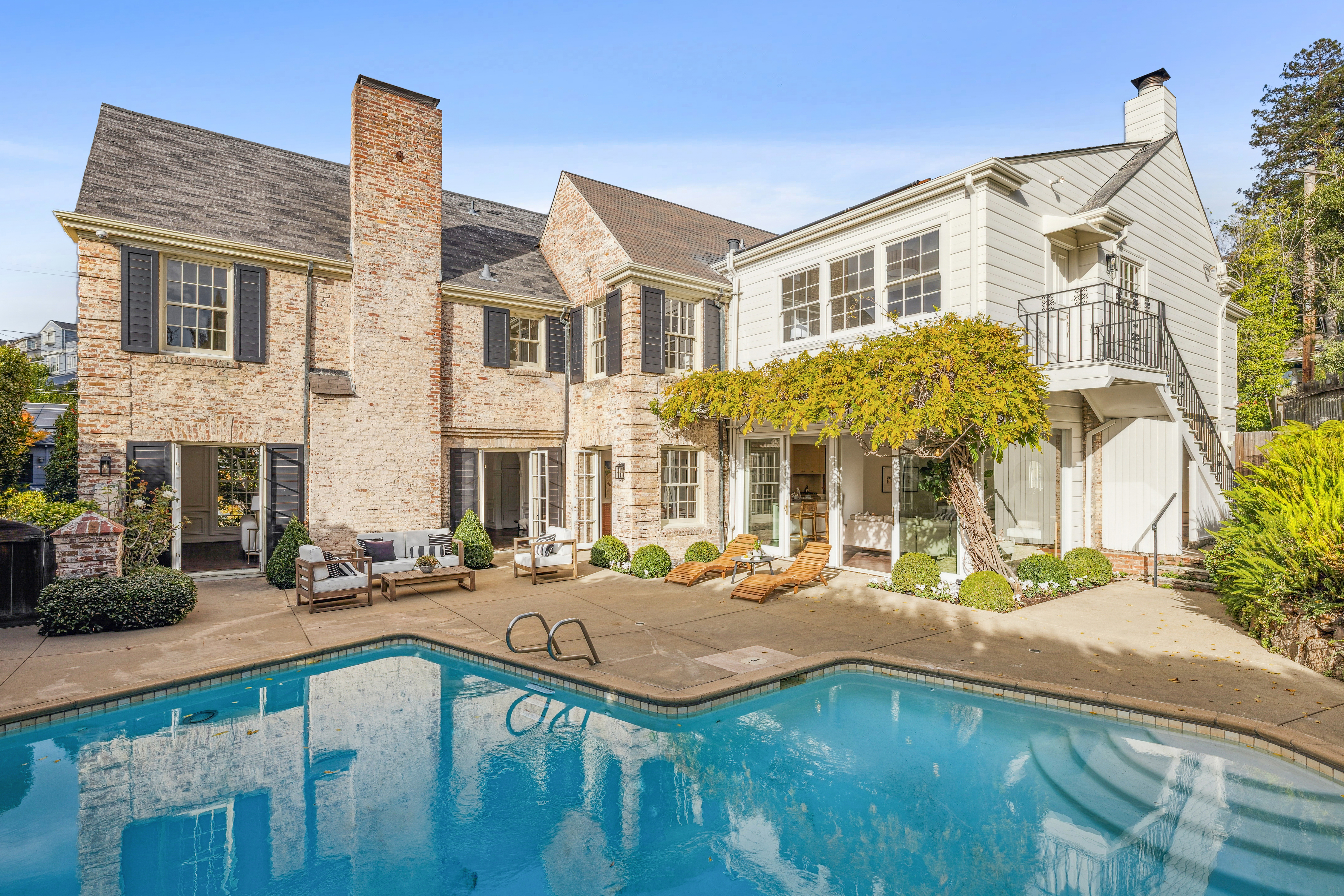 This image showcases the rear exterior of a multi-story brick and white-painted home, highlighting a pristine swimming pool as the focal point. The pool is surrounded by a concrete patio featuring lounge chairs and outdoor seating, creating an inviting space for relaxation and entertainment. The architecture blends classic brick elements with modern white siding, complemented by lush landscaping and a mature tree providing shade.