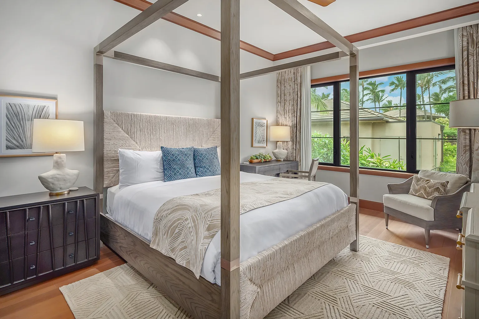 This is a well-appointed primary bedroom featuring a four-poster bed with a woven headboard and base, complemented by blue accent pillows and a patterned throw. A dark wood nightstand with a modern lamp sits beside the bed, while a comfortable armchair is positioned near the window, which offers a view of lush greenery. The room is finished with a geometric patterned rug, creating a serene and inviting atmosphere.