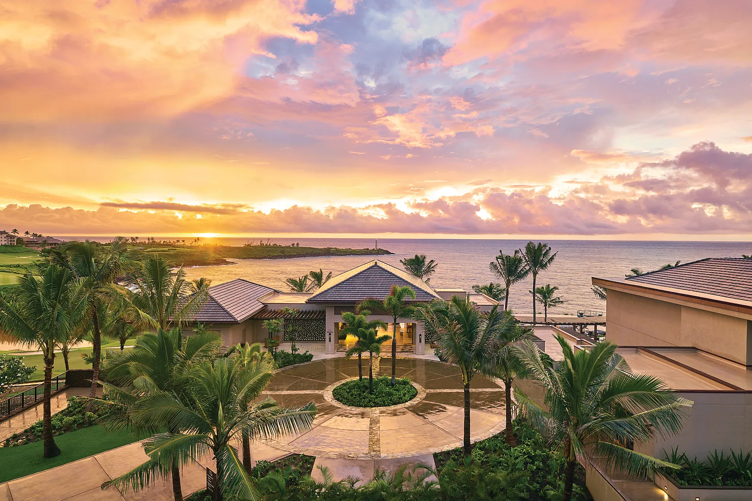 This aerial view showcases a luxurious beachfront property at sunset, featuring a grand entrance with a circular driveway and palm trees. The building's architecture blends seamlessly with the tropical landscape, offering stunning ocean views and a sense of exclusivity. The overall impression is one of opulence and tranquility.