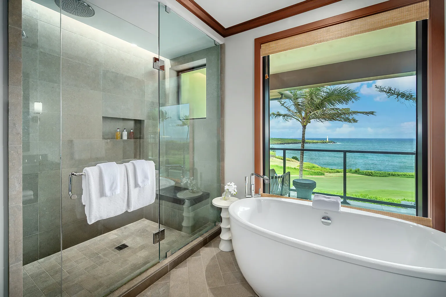 This is a luxurious primary bathroom featuring a glass-enclosed shower with a rainfall showerhead and built-in niche, a freestanding soaking tub positioned in front of a large window with ocean views, and neutral-toned tile flooring. The bathroom exudes a spa-like ambiance with its clean lines, natural light, and serene color palette, creating a relaxing and upscale experience. The view from the window includes a palm tree, the ocean, and a lighthouse in the distance.