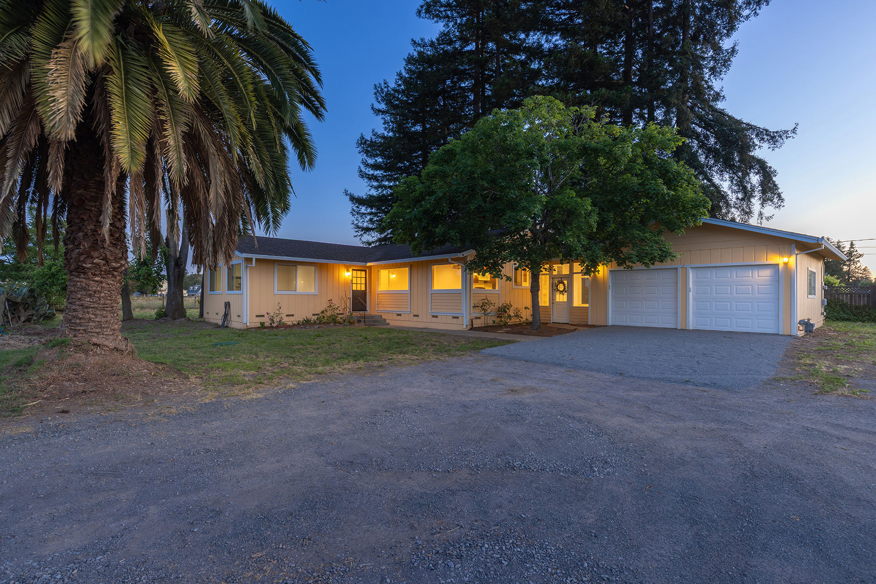 This single-story, yellow-sided home is captured at dusk, showcasing a warm and inviting atmosphere with illuminated windows and exterior lighting. The property features a prominent palm tree in the foreground, a gravel driveway leading to a two-car garage, and a mature tree shading the front entrance. The perspective is a wide-angle shot from the driveway, emphasizing the home's sprawling layout and its integration with the surrounding landscape.