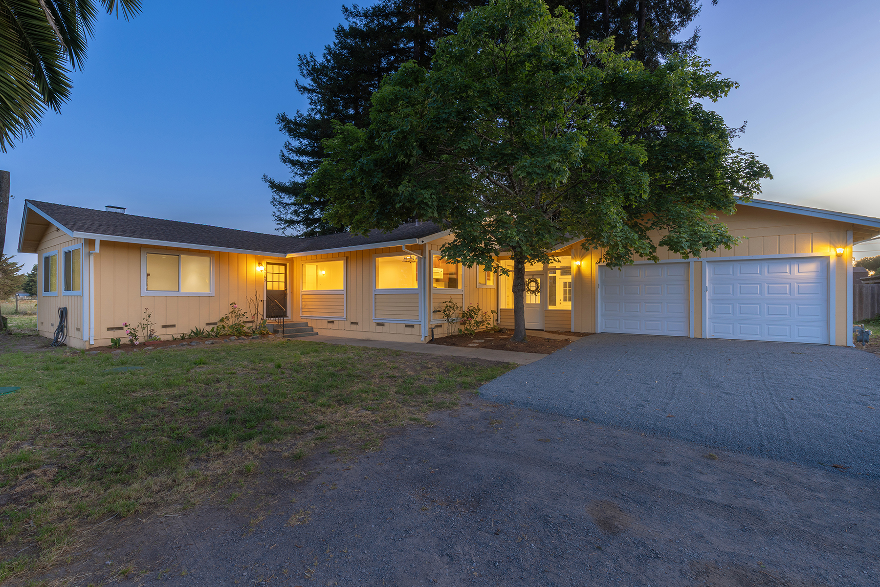 This single-story, yellow-sided ranch-style home is captured at dusk, showcasing a warm and inviting atmosphere with interior lights glowing through the windows. The front facade features a multi-car garage, a central entryway, and a large, mature tree centered in the front yard. A gravel driveway leads up to the garage, while the surrounding landscape includes a mix of lawn and natural shrubbery under a clear twilight sky.