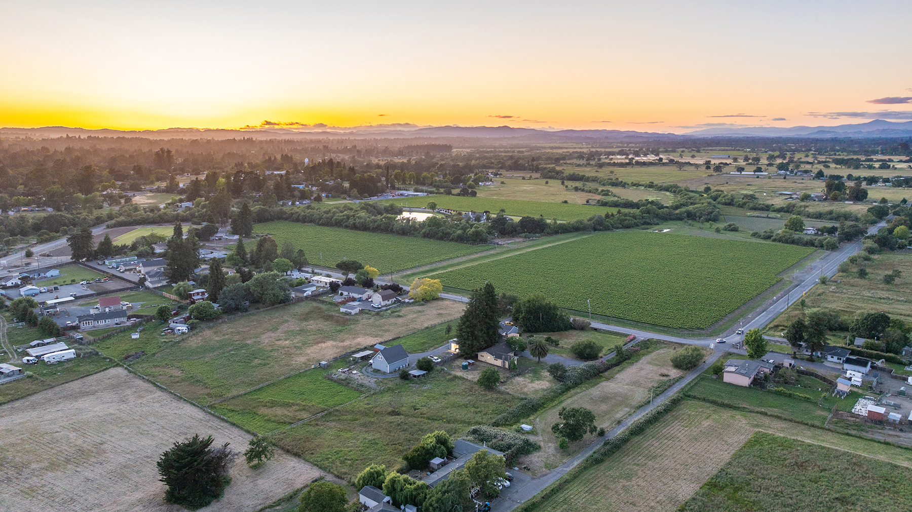 This expansive aerial view captures a rural landscape at sunset, showcasing a mix of residential properties, open fields, and large agricultural vineyards. The scene is bathed in a warm, golden light, highlighting the rolling hills in the distance and the quiet, sprawling nature of the countryside. The perspective provides a comprehensive look at the property's surroundings, emphasizing its peaceful setting and proximity to open land.