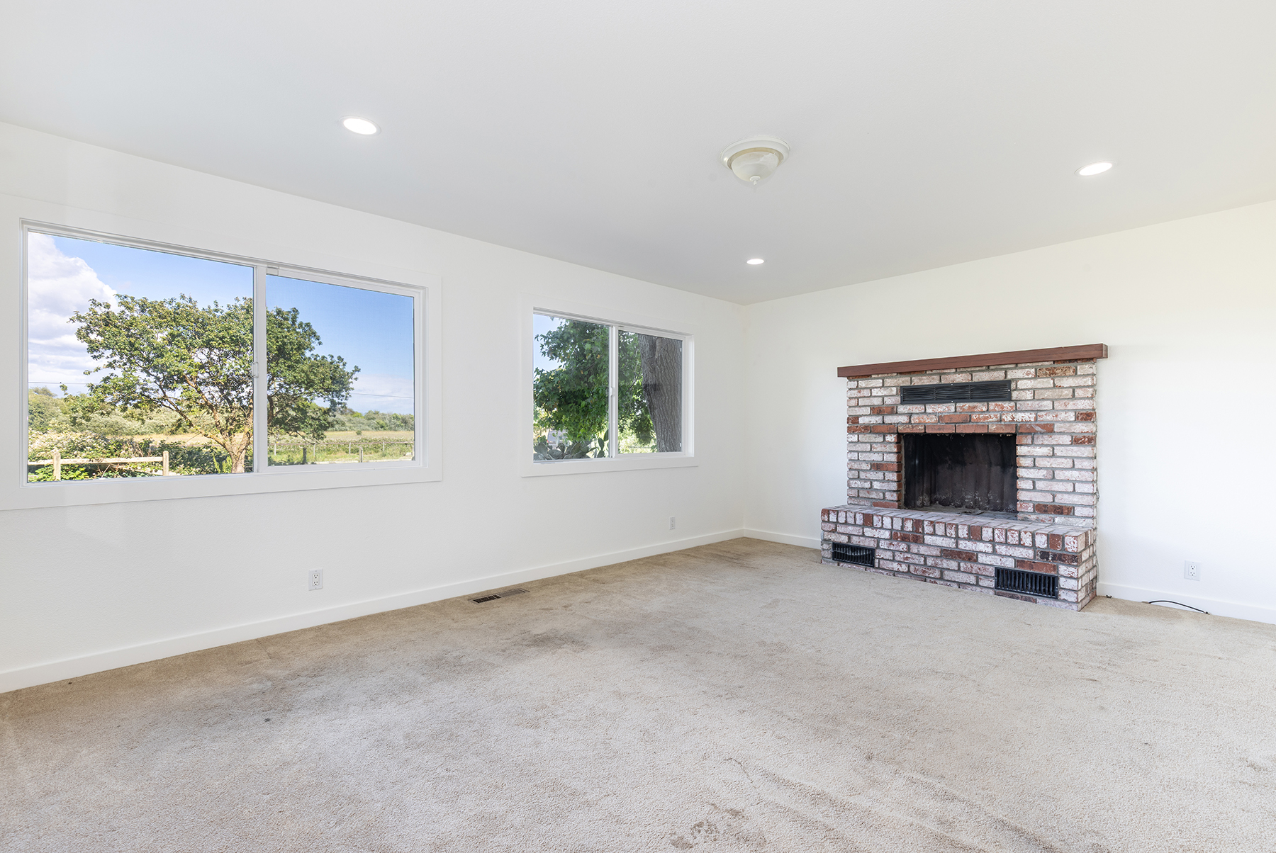This bright living room features neutral walls and wall-to-wall carpeting, providing a blank canvas for personalization. A prominent brick fireplace with a wood mantel serves as the focal point, while two large windows offer expansive views of the surrounding landscape. The room is illuminated by recessed lighting and a central ceiling fixture, creating a clean and open atmosphere.