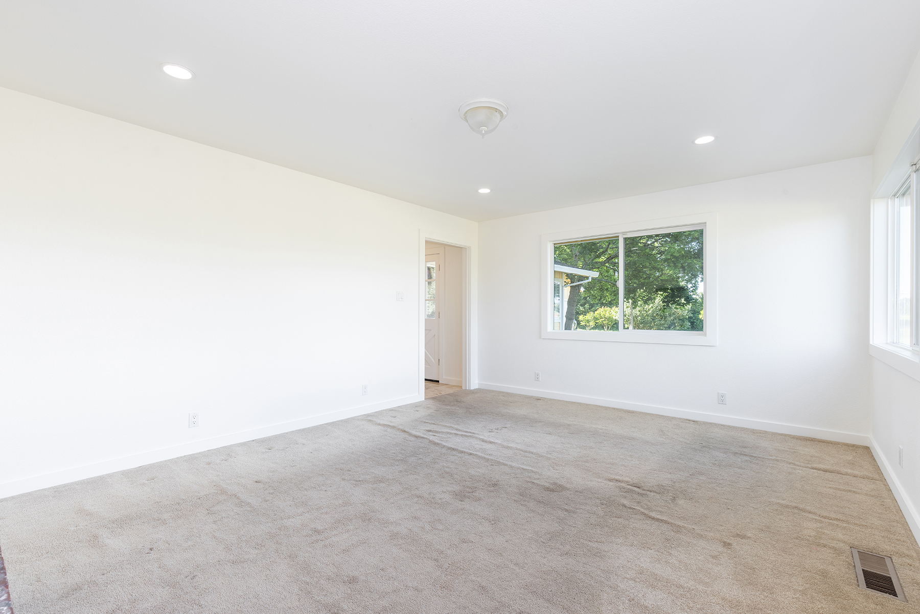 This bright and neutral room features plush carpeting and crisp white walls, creating a clean, blank canvas ready for personalization. A large window allows for ample natural light, while recessed lighting and a central ceiling fixture provide additional illumination. The space includes a doorway leading to an adjacent area, offering a functional layout suitable for a bedroom or home office.
