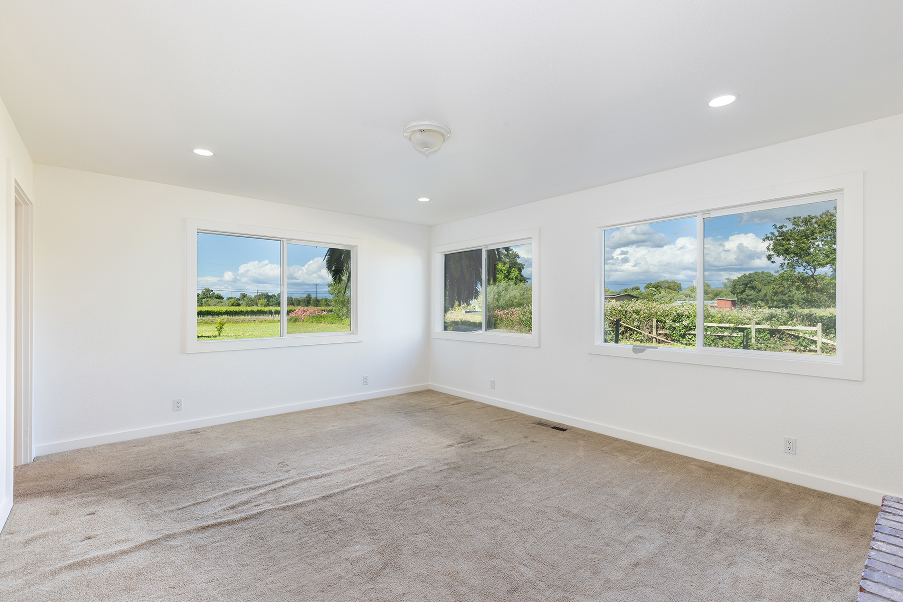 This bright and spacious room features neutral white walls and wall-to-wall carpeting, providing a clean canvas for any design style. Three large windows offer abundant natural light and expansive views of the surrounding rural landscape. The room is illuminated by recessed lighting and a central ceiling fixture, creating an open and airy atmosphere.
