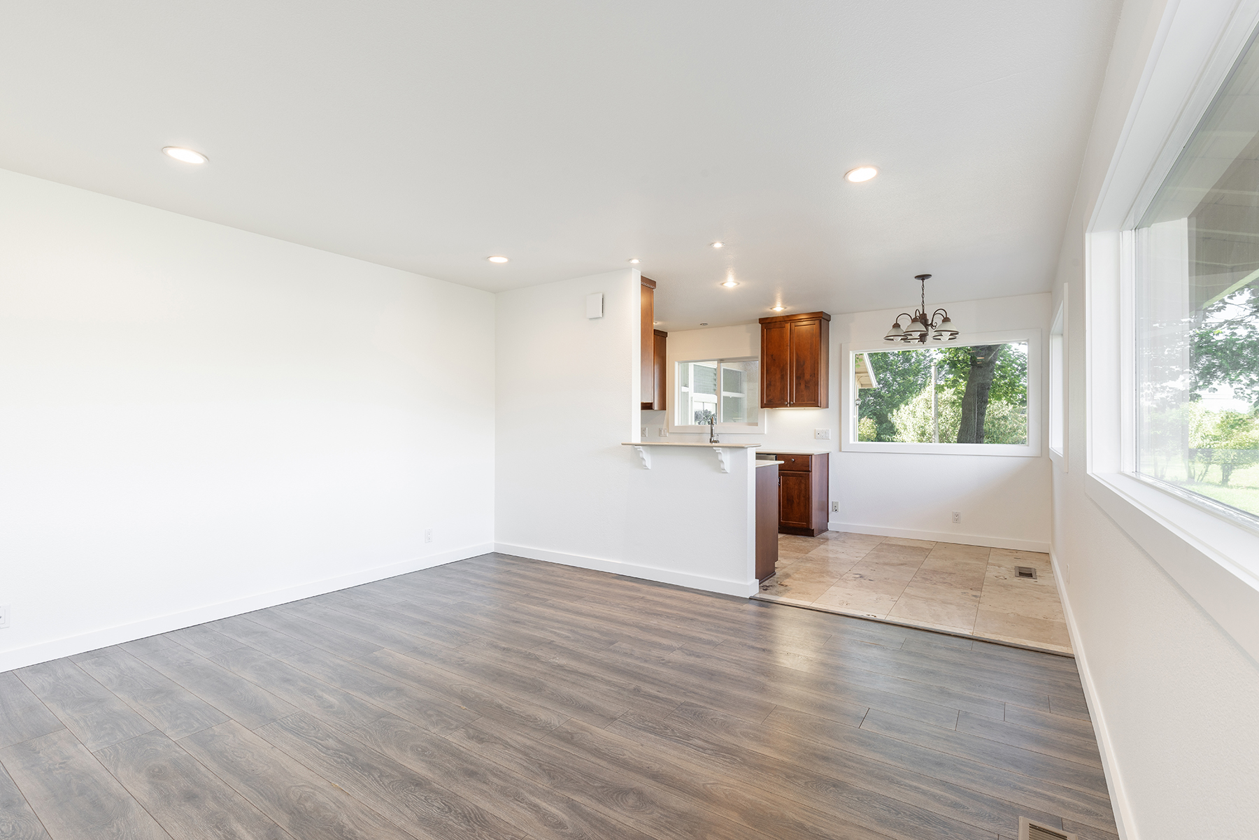 This open-concept living area features dark wood-look flooring that transitions into a tiled dining space near the kitchen. The room is bright and neutral, with a breakfast bar peninsula separating the living area from the kitchen, which is equipped with dark wood cabinetry. Large windows provide ample natural light and views of the outdoors, creating an airy and inviting atmosphere.