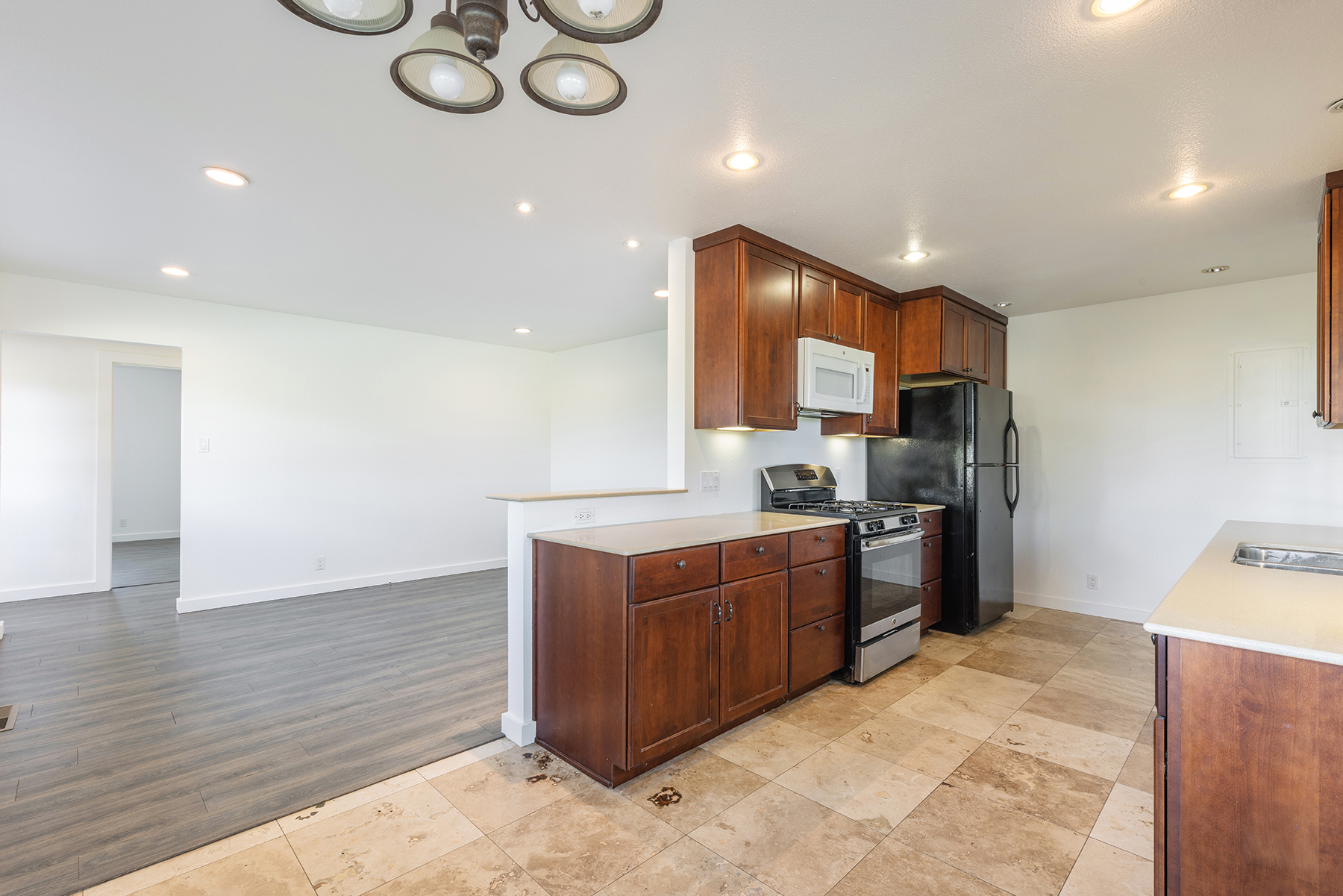 This kitchen features warm-toned wood cabinetry paired with light-colored countertops and a black refrigerator and stove. The space is open to an adjacent living area with dark wood-look flooring, while the kitchen itself is finished with large, neutral-toned stone floor tiles. Recessed lighting and a decorative chandelier provide illumination, creating a functional and inviting atmosphere.