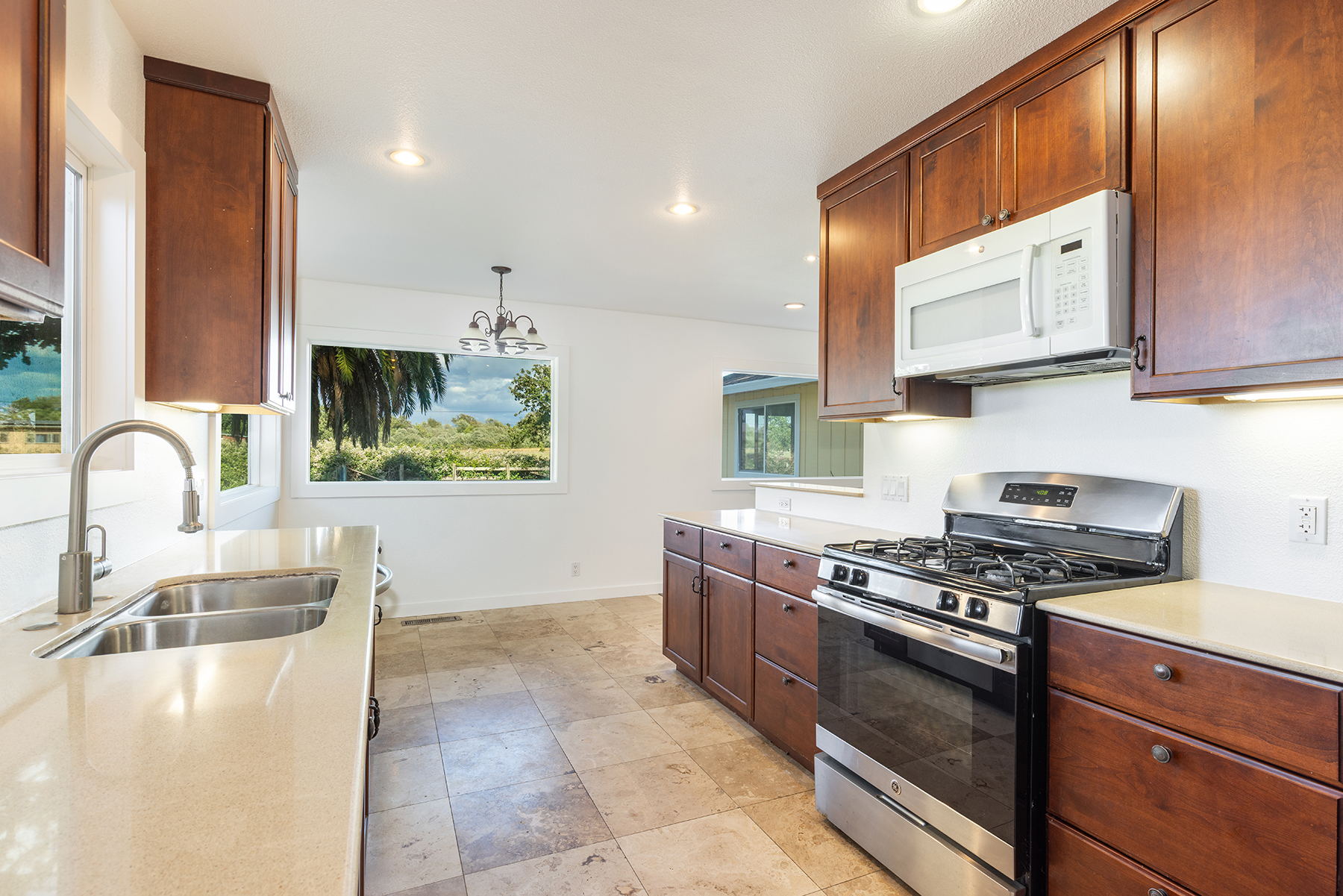This bright, well-lit kitchen features warm-toned wood cabinetry paired with light-colored countertops and a stainless steel gas range. The space is anchored by a double-basin sink under a window and a large picture window that provides a scenic view of the outdoors. The neutral-toned tile flooring and recessed lighting create a clean, functional, and inviting atmosphere.