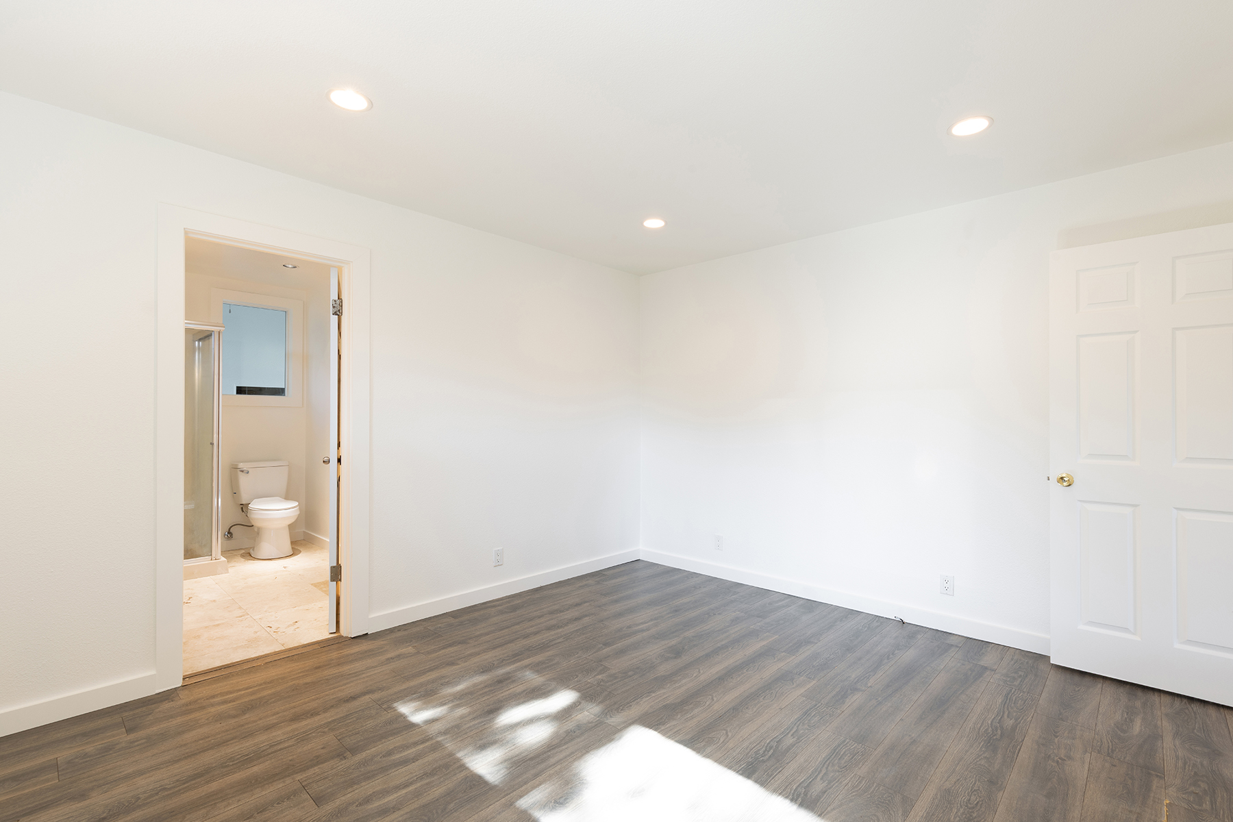 This bright, minimalist bedroom features neutral white walls and dark wood-look flooring, creating a clean and versatile living space. A doorway on the left provides a direct view into an attached bathroom, while a white paneled door on the right leads to another area of the home. The room is illuminated by recessed ceiling lights, offering a simple and modern aesthetic suitable for a guest room or home office.