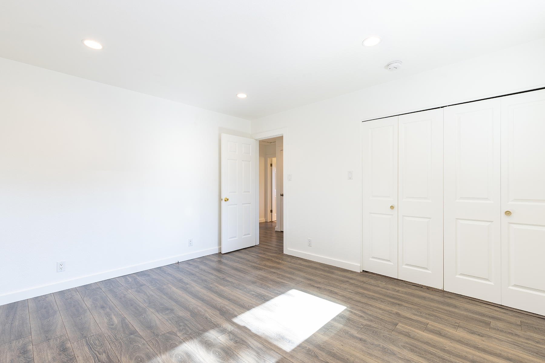 This bright, minimalist bedroom features neutral white walls and modern wood-look flooring. The room includes a wide closet with double doors, a doorway leading to a hallway, and recessed lighting throughout. The clean, open layout provides a versatile space ready for personalization.