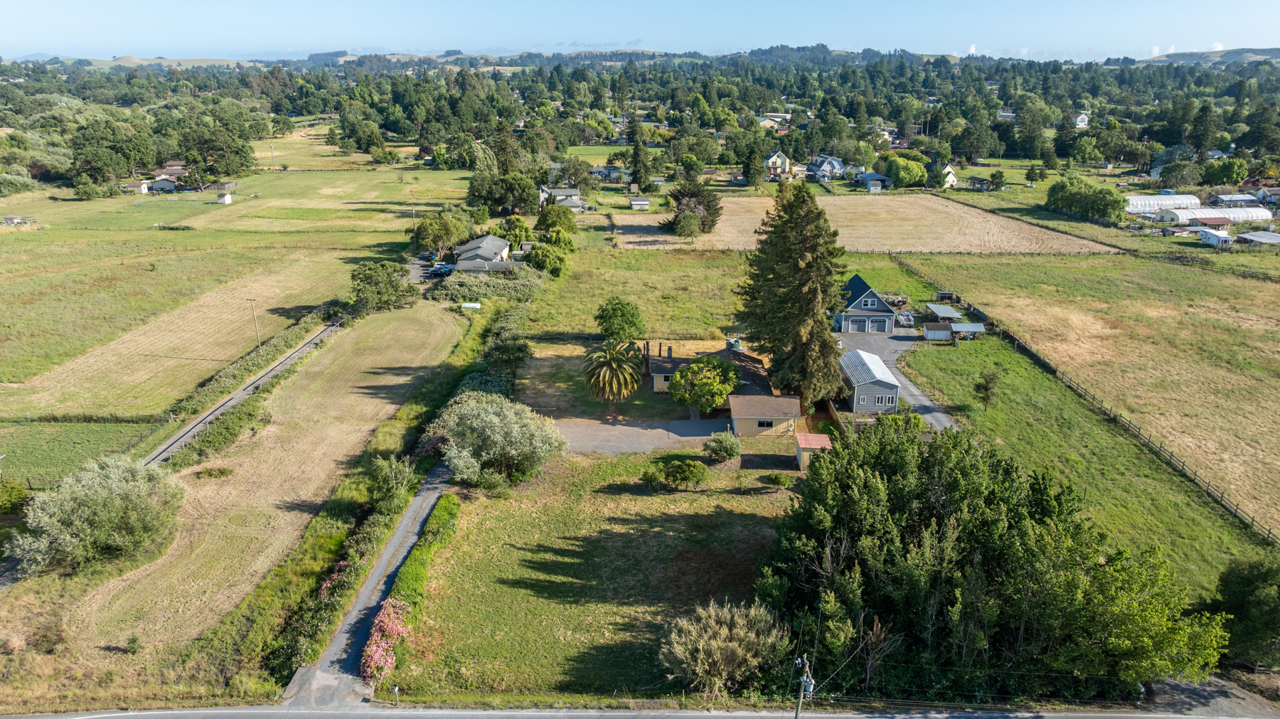 This high-angle aerial view captures a sprawling rural property set amidst expansive green fields and mature trees. The main residence, accompanied by a detached garage and secondary outbuildings, is situated on a large, open lot with a long driveway leading from the road. The surrounding landscape features rolling hills in the distance, emphasizing the quiet, private nature of this countryside estate.