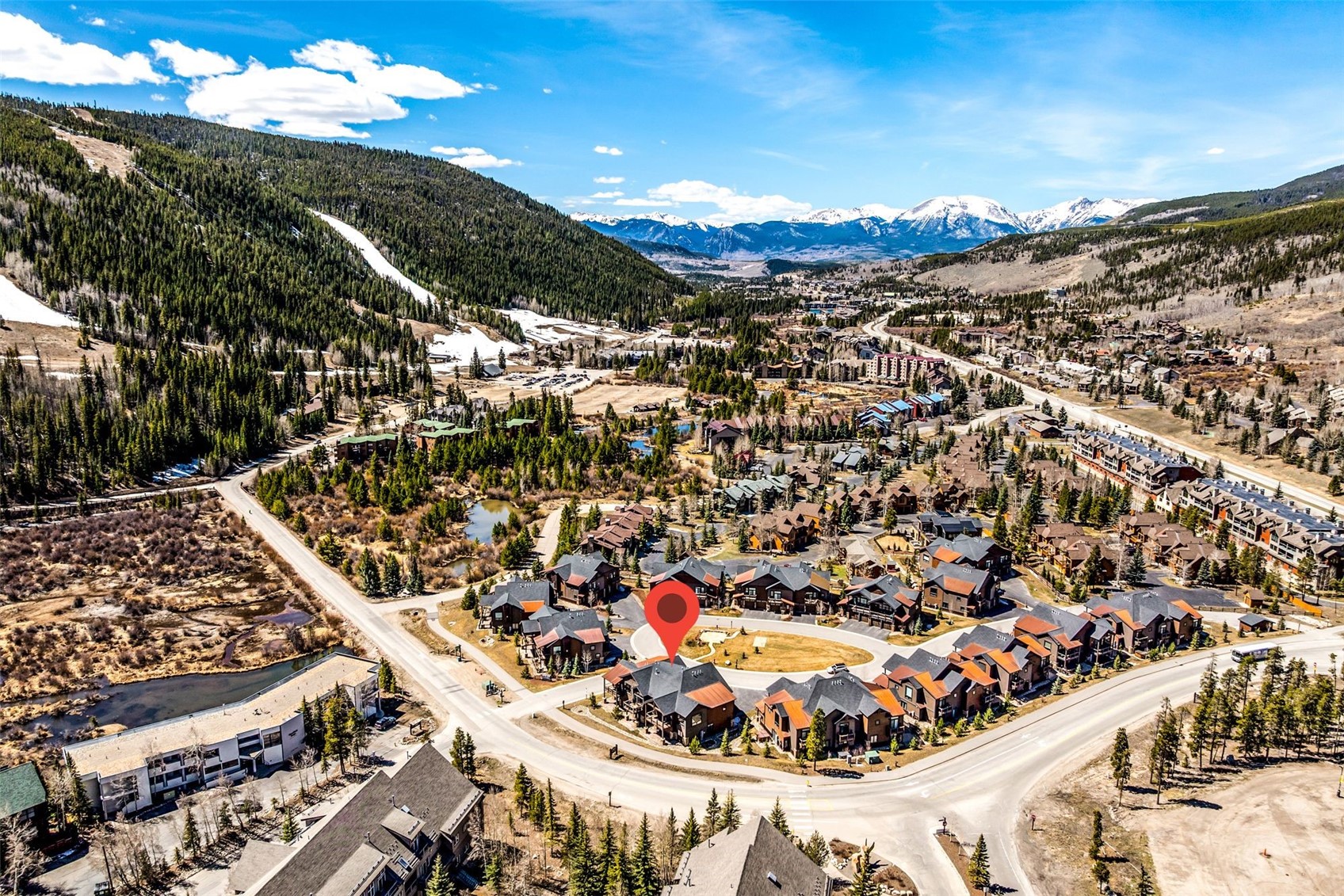 This high-angle, aerial perspective displays a residential alpine complex situated in a mountainous landscape, marked by a prominent red location pin. The neighborhood features distinctive architectural styles with dark, multi-faceted rooflines, blending seamlessly into the expansive valley floor and surrounding pine forests. The scenic backdrop contains snow-capped mountain peaks, emphasizing a premier location ideal for mountain recreational life.
