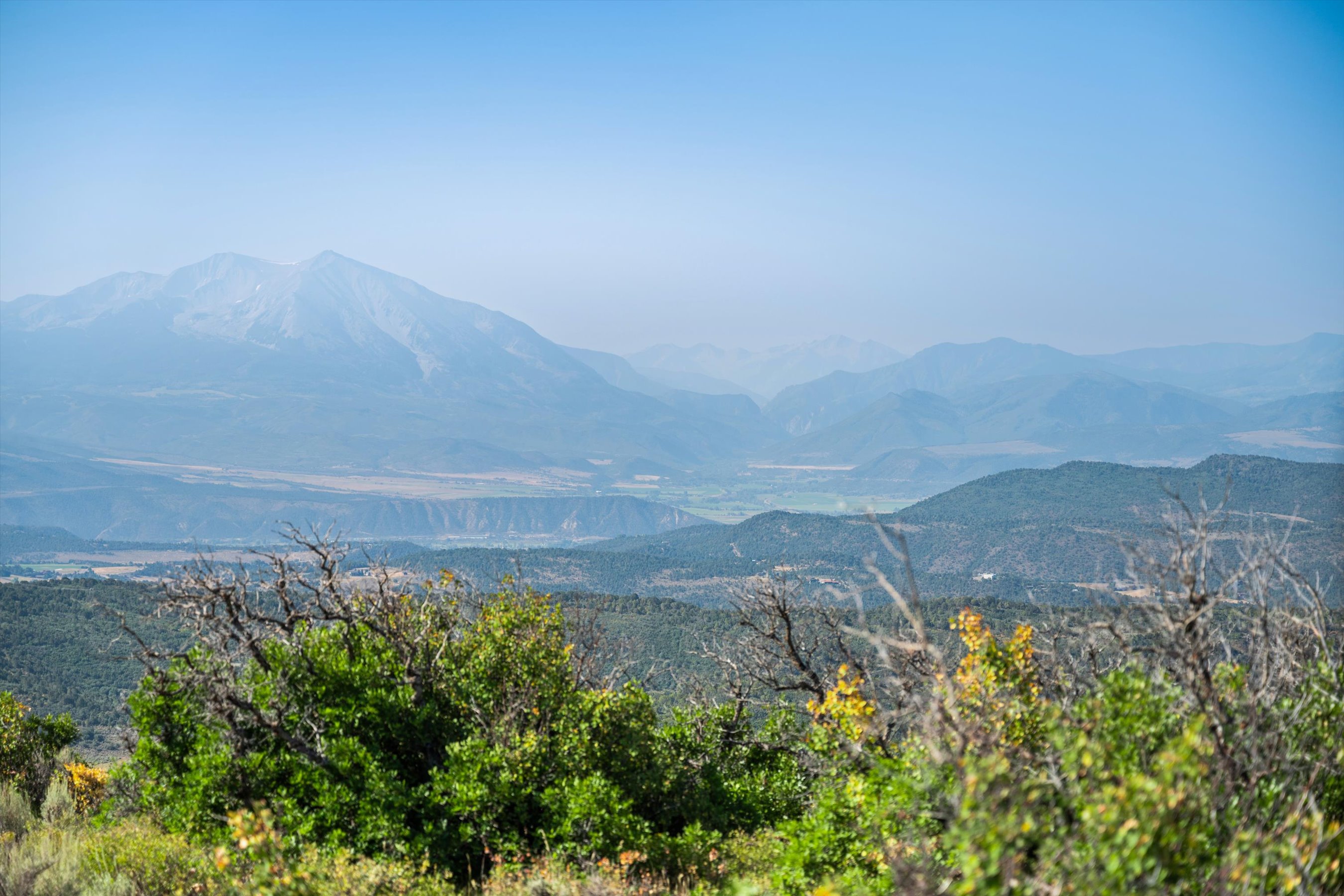 This image captures a breathtaking, expansive landscape view of a mountain range under a clear, bright blue sky. In the foreground, lush green shrubs and dry, skeletal branches frame the scene, providing a natural depth that leads the eye toward the valley and the towering, hazy peaks in the distance. The perspective is elevated, suggesting a scenic overlook or a vantage point from a high-altitude property, emphasizing the serene and majestic natural surroundings.
