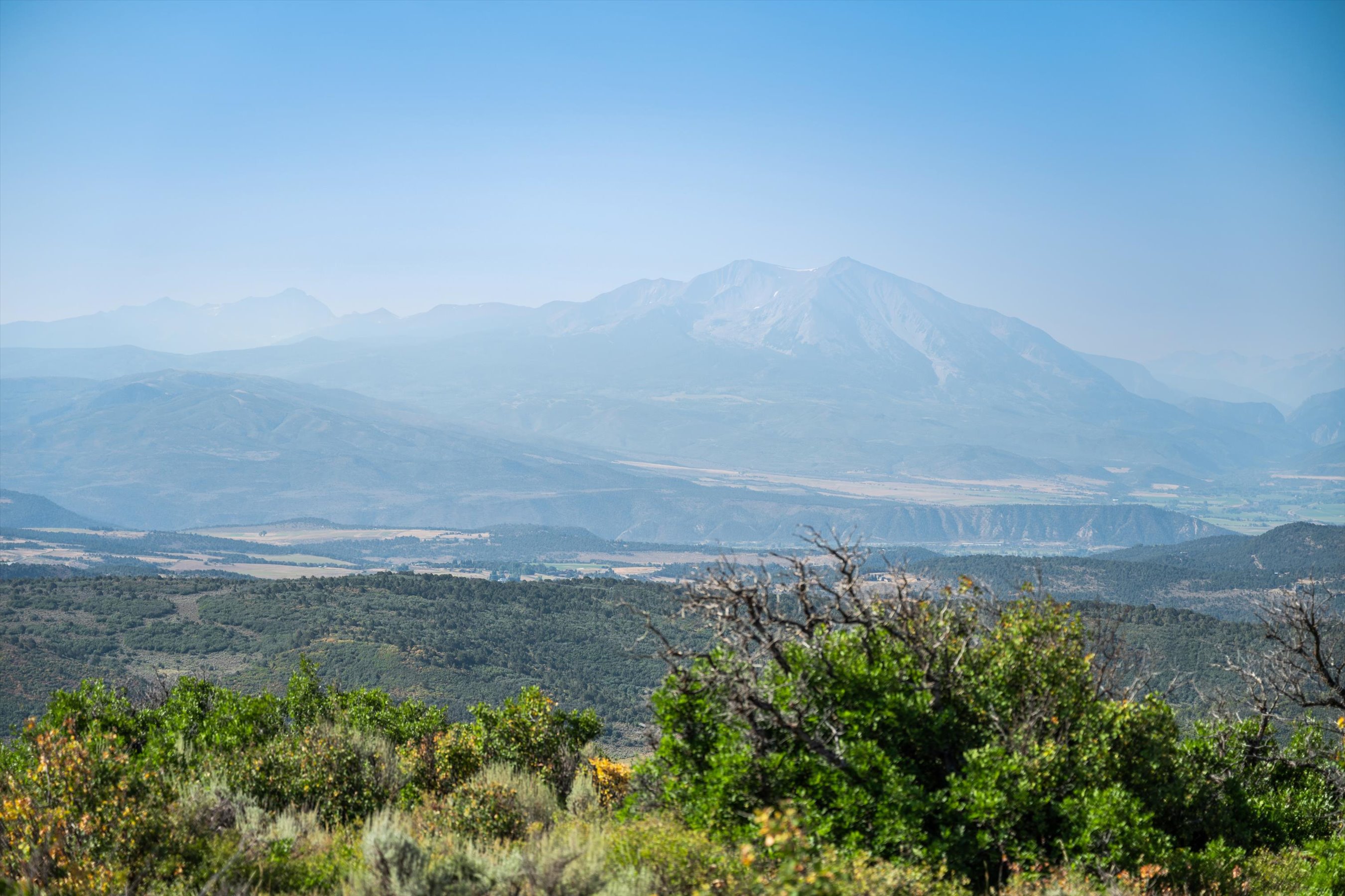 This scenic landscape shot captures a vast, hazy mountain range under a clear blue sky, viewed from an elevated vantage point. In the foreground, lush green shrubs and trees frame the expansive valley below, which features rolling hills and open fields. The perspective is wide and cinematic, emphasizing the natural beauty and serene atmosphere of the property's surroundings.