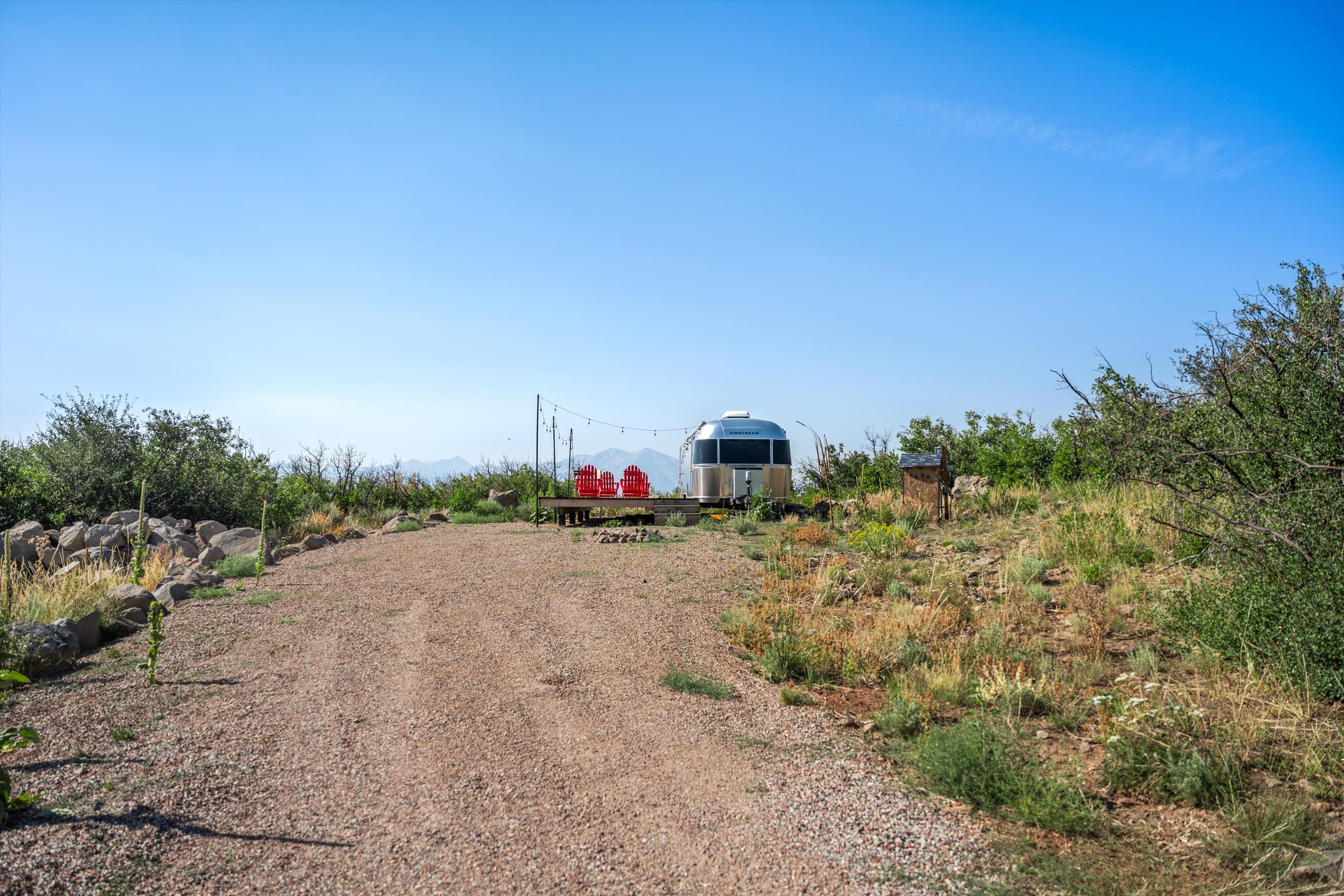 This wide-angle shot captures a scenic, elevated campsite featuring a classic silver Airstream trailer parked on a gravel lot. A wooden deck with vibrant red Adirondack chairs provides a perfect vantage point for the distant mountain range under a clear blue sky. The surrounding landscape is rugged and natural, emphasizing a peaceful, off-grid retreat atmosphere.